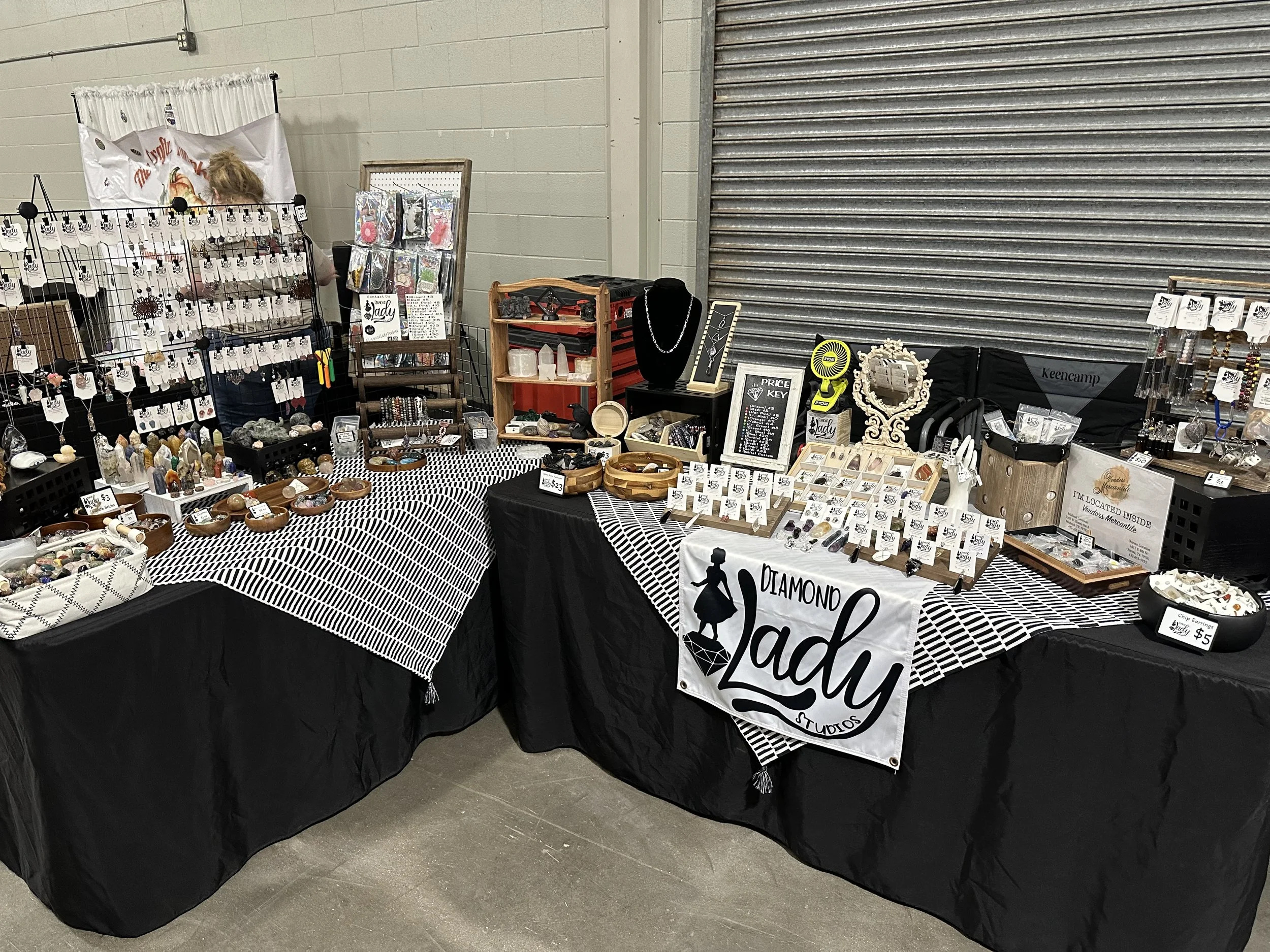 Jewelry booth at a market with necklaces, bracelets, and stones on display, with a black and white checkered tablecloth and a sign that reads 'Diamond Lady''. A person with blonde hair is behind the booth, partially obscured by display racks.