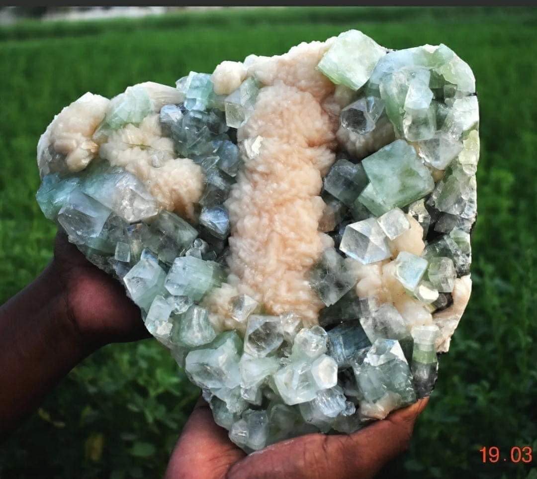 A person's hands holding a large mineral or crystal formation with green, white, and gray hues, set against a green outdoor background.