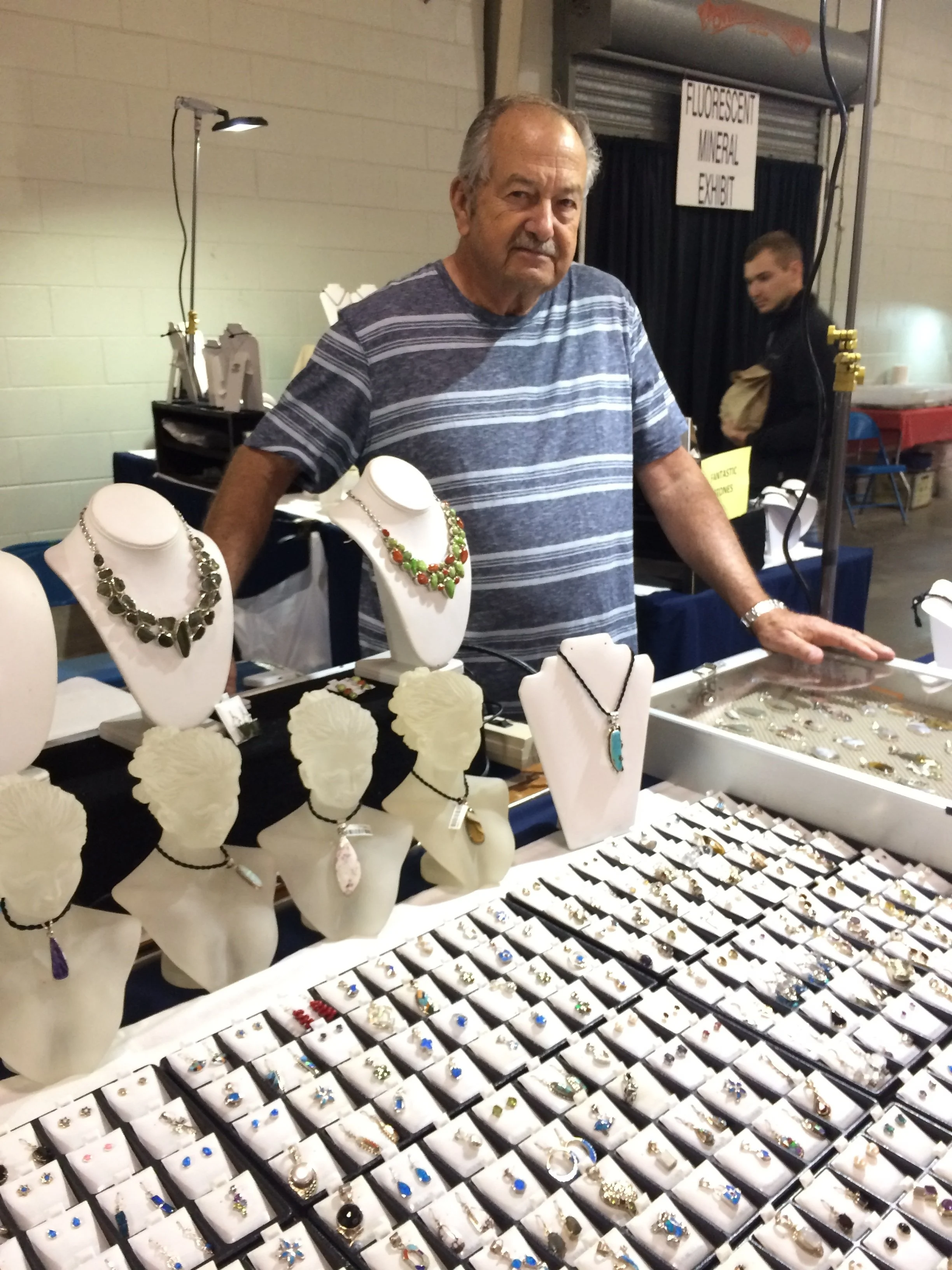 An older man standing behind a jewelry display at a market or fair. The display has necklaces and rings, some with colorful stones or charms. In the background, another person is holding a paper bag near a sign that reads 'Fluorescent Mineral Exam'.