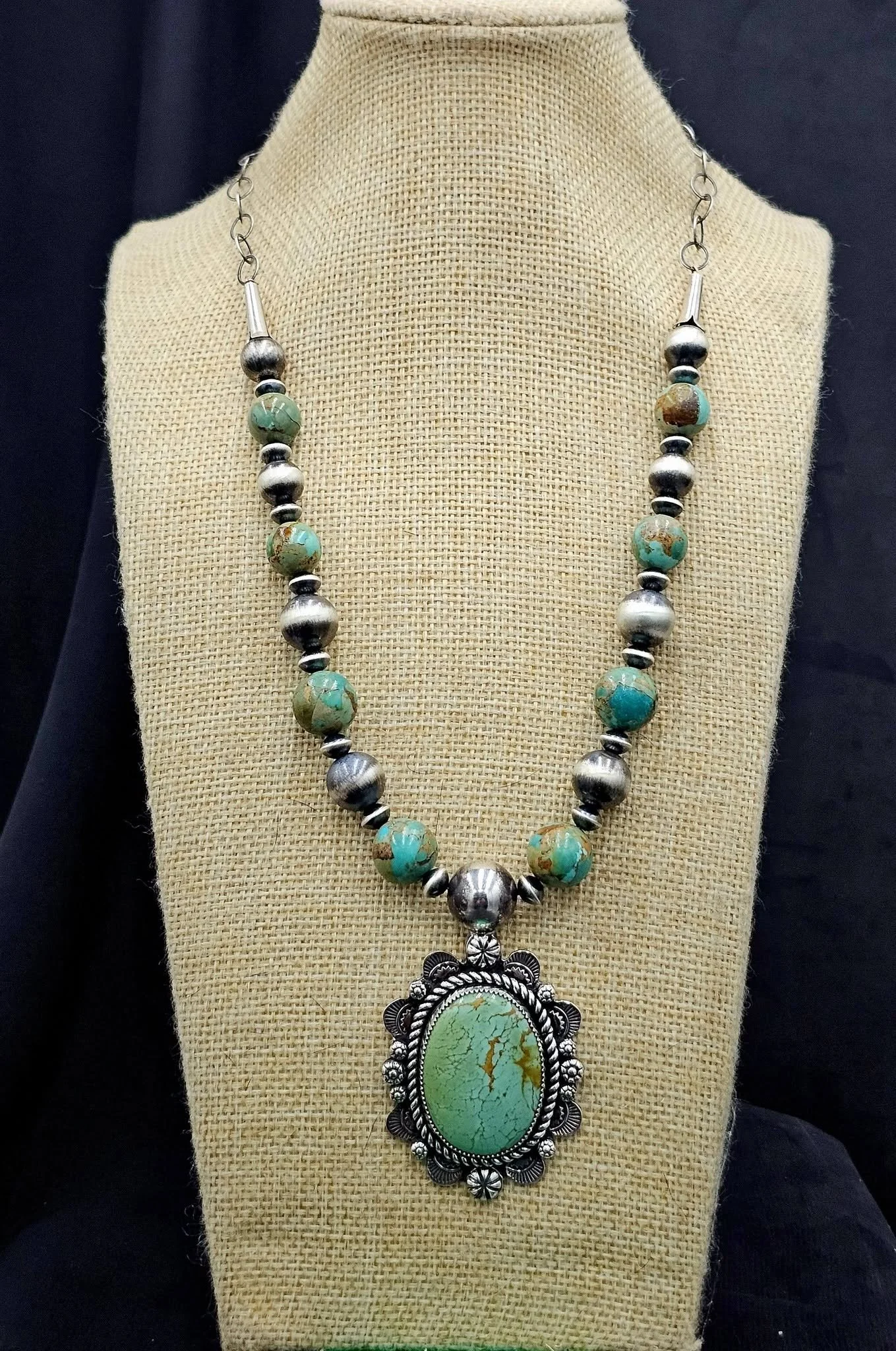 A turquoise and silver beaded necklace with a large ornate pendant featuring a green turquoise stone, displayed on a beige fabric bust.