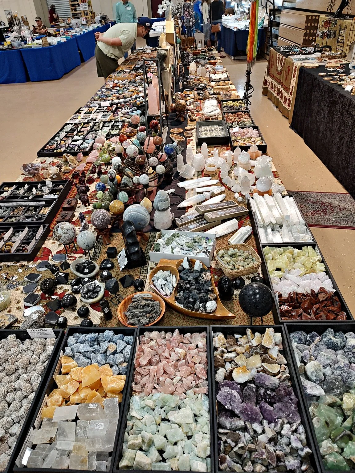 A marketplace stall with a variety of gemstones, minerals, and crystals. Displayed on tables and in boxes, including polished stones, rough minerals, and decorative items. People browsing in the background.