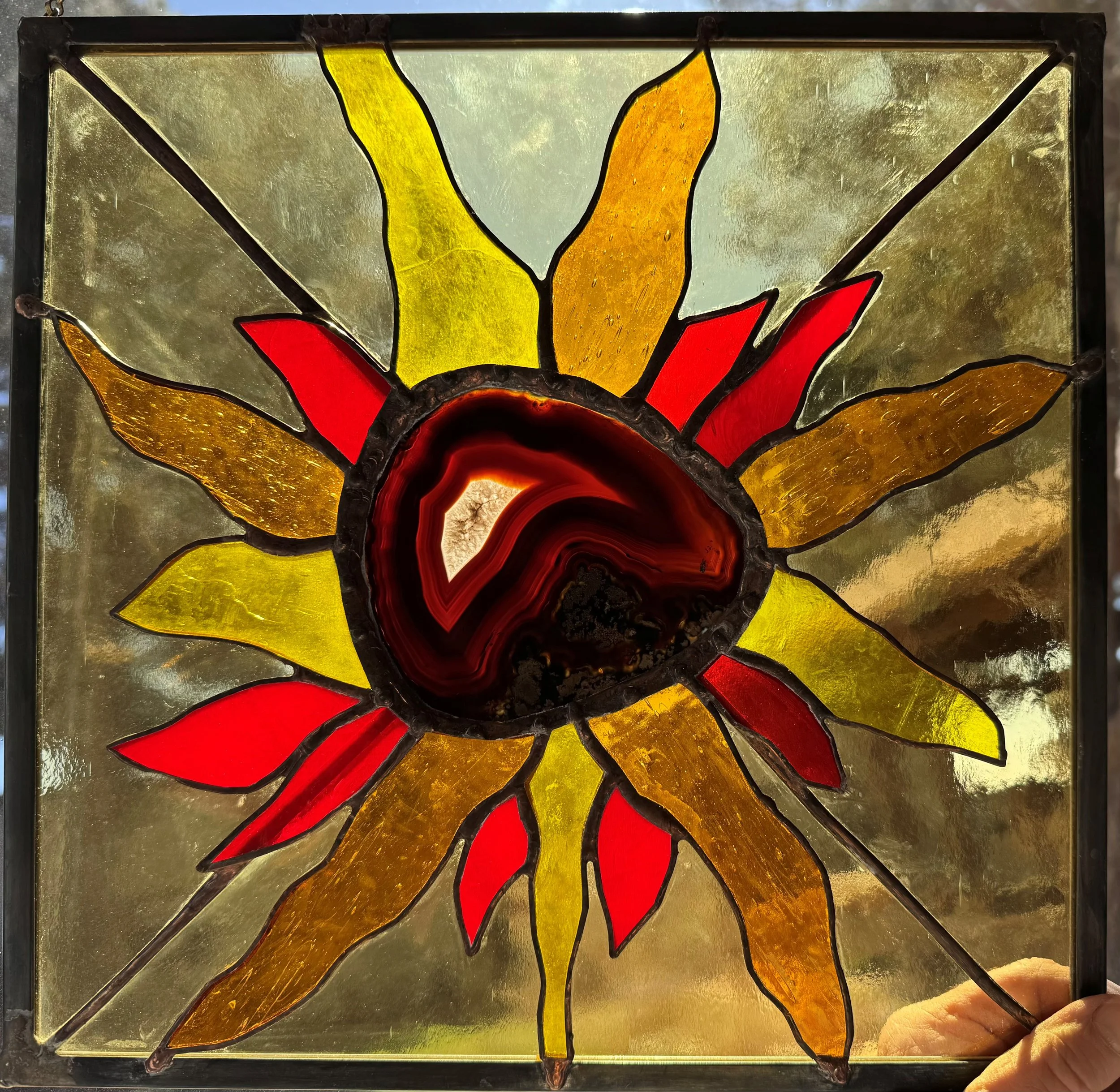 A stained glass window depicting a stylized sunflower with yellow and orange petals and a red center, set within a black frame.