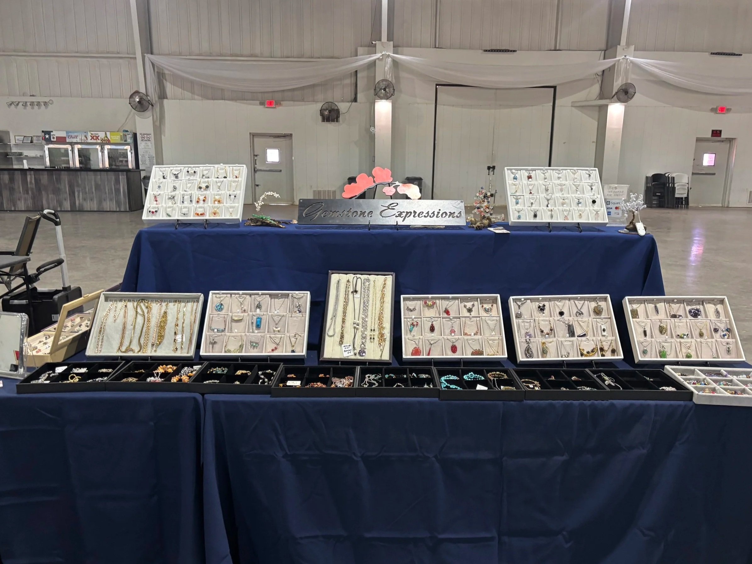 Jewelry display tables at an indoor market or fair, showcasing rings, necklaces, and earrings, with a sign that reads 'Garnstone Expressions'.