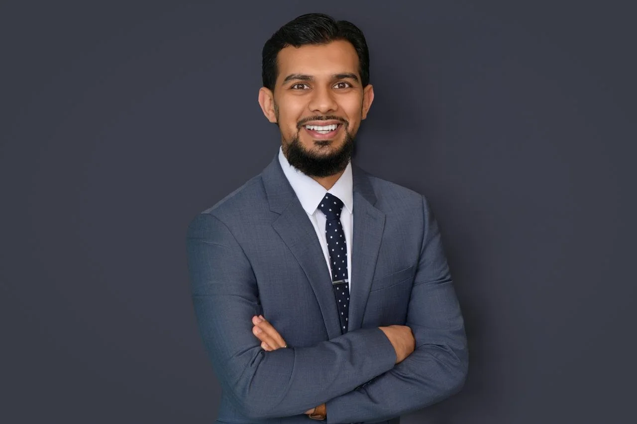 Professional man in a grey suit with a white shirt and polka dot tie smiling against a dark background.