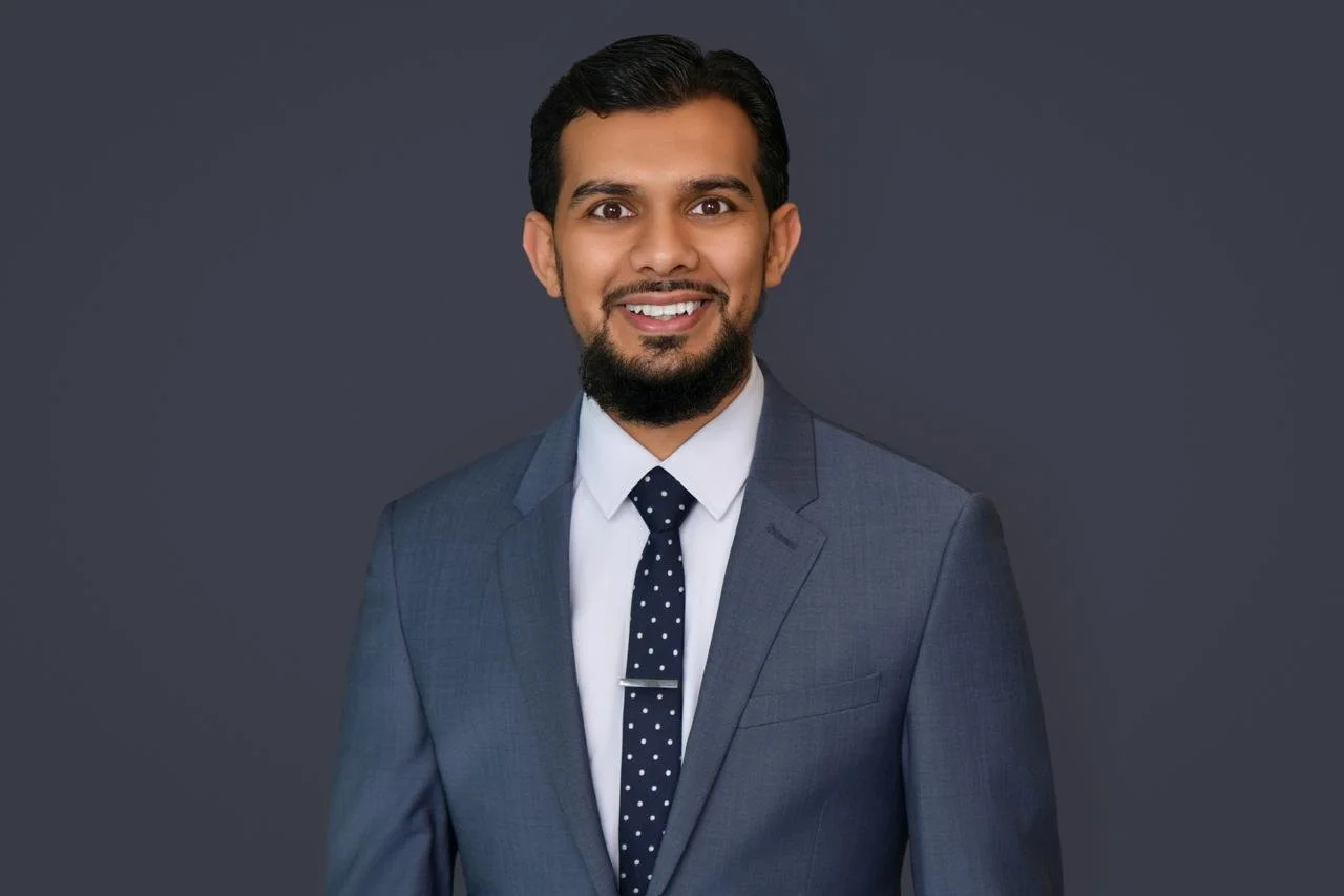 Professional headshot of a man in a gray suit, white shirt, and navy polka dot tie, smiling against a dark gray background.