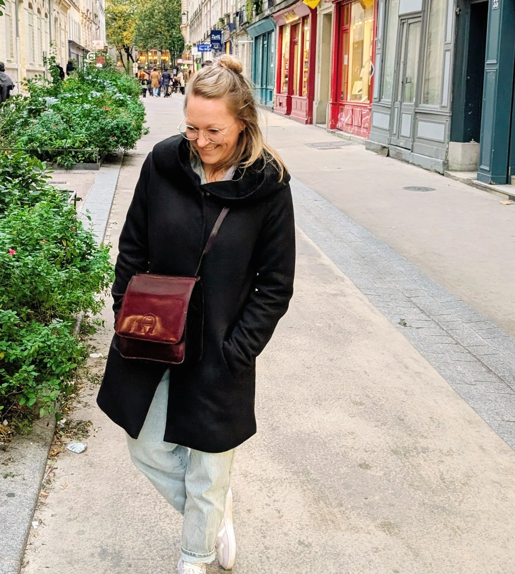 Eine lachende Frau mit Brille trägt eine schwarze Jacke, eine weiße Hose und weiße Turnschuhe, während sie auf einer belebten Straße in einer Stadt spazieren geht. Straße in Paris nach Flohmarktbesuch.