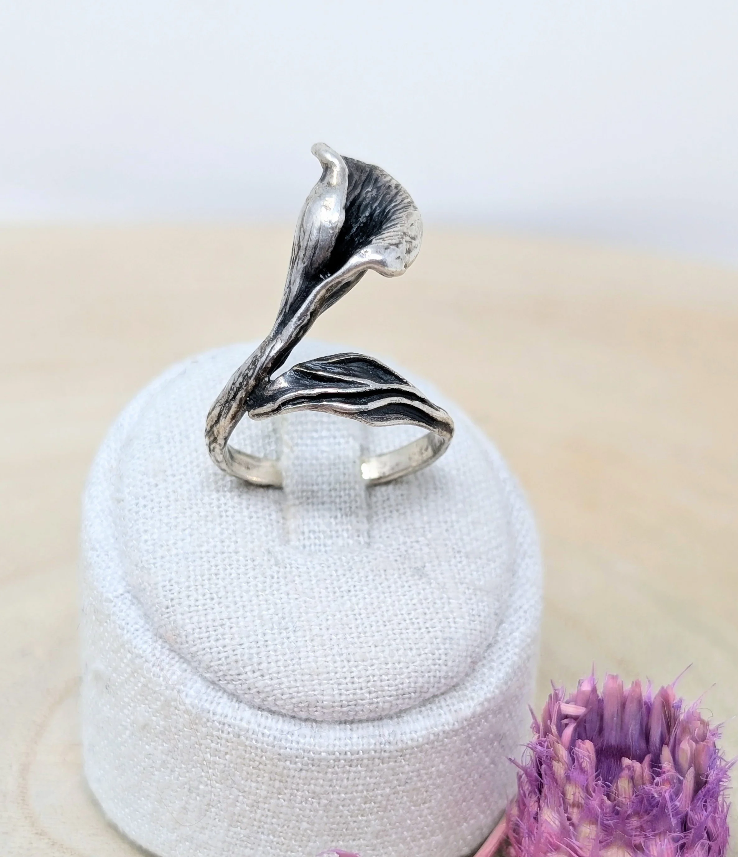 Silberner Ring in Form einer Blume auf einer weißen, texturierten Unterlage, mit lila Blüten im Vordergrund. Besonderer 925 silber Ring vintage, Juwelier-Arbeit