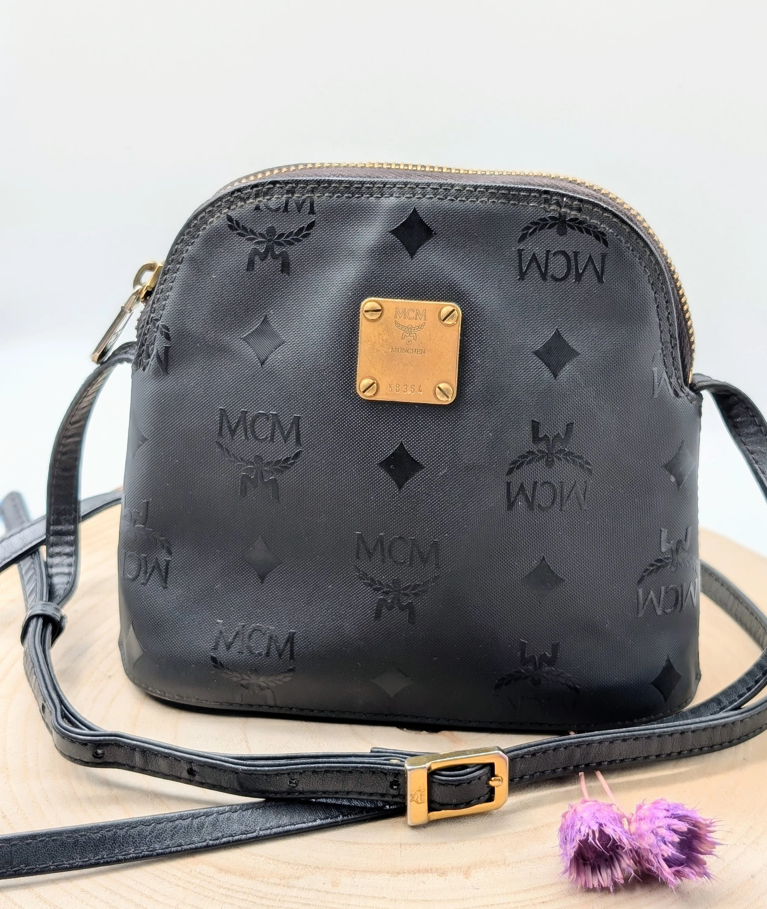 Schwarze Tasche mit Goldakzenten und MCM Logo-Muster, auf einem Holztablett mit einem schwarzen Riemen und einer goldfarbenen Schnalle, neben rosa- und lila-farbene Pompons. Vintage Michael Cromer München