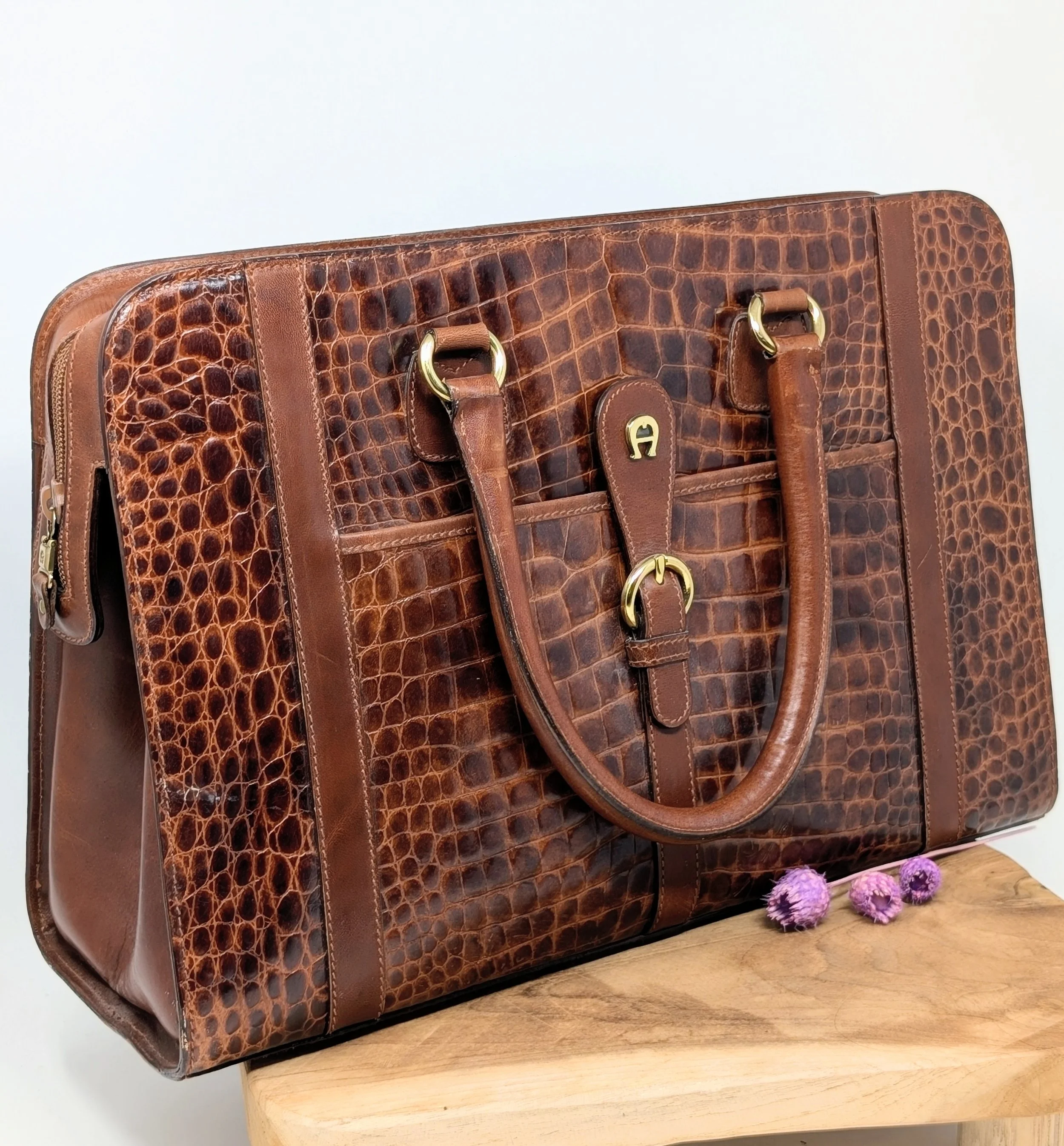 Eine braune Ledertasche mit Krokodil Muster, goldenen Schnallen und mehreren Fächern, liegt auf einem Holz-Tisch, mit kleinen lila und rosa Blumen daneben. Marke Aigner, Etienne Aigner, Vintage