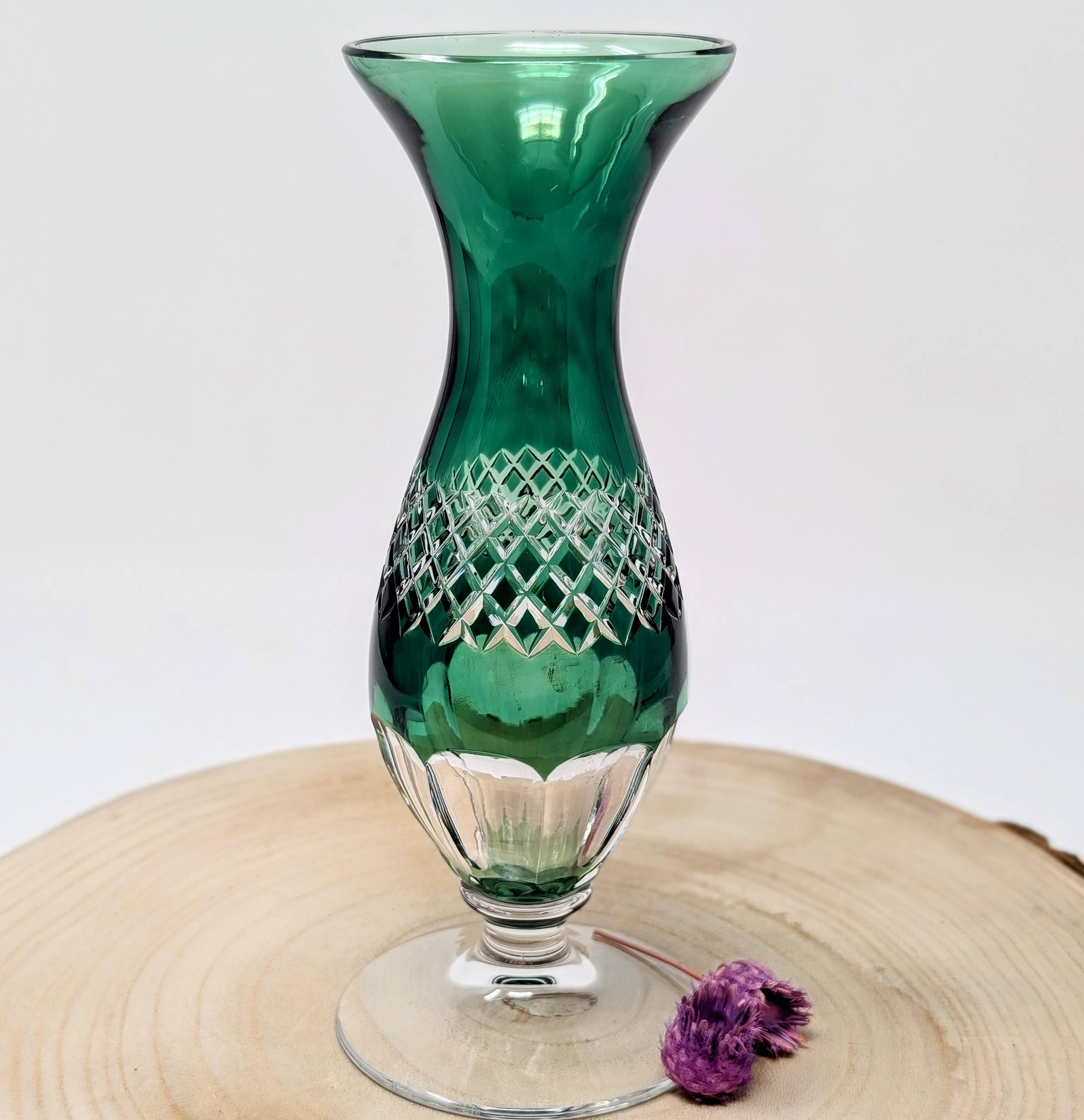 Ein grünes, kunstvoll gestaltetes Glas mit geschnitztem Muster am unteren Rand, das auf einem runden Holztablett steht, mit vier kleinen lila Blütenblättern oder Blumen auf der Oberfläche. Lalique Vase, Kristallglas, Überfang grün.