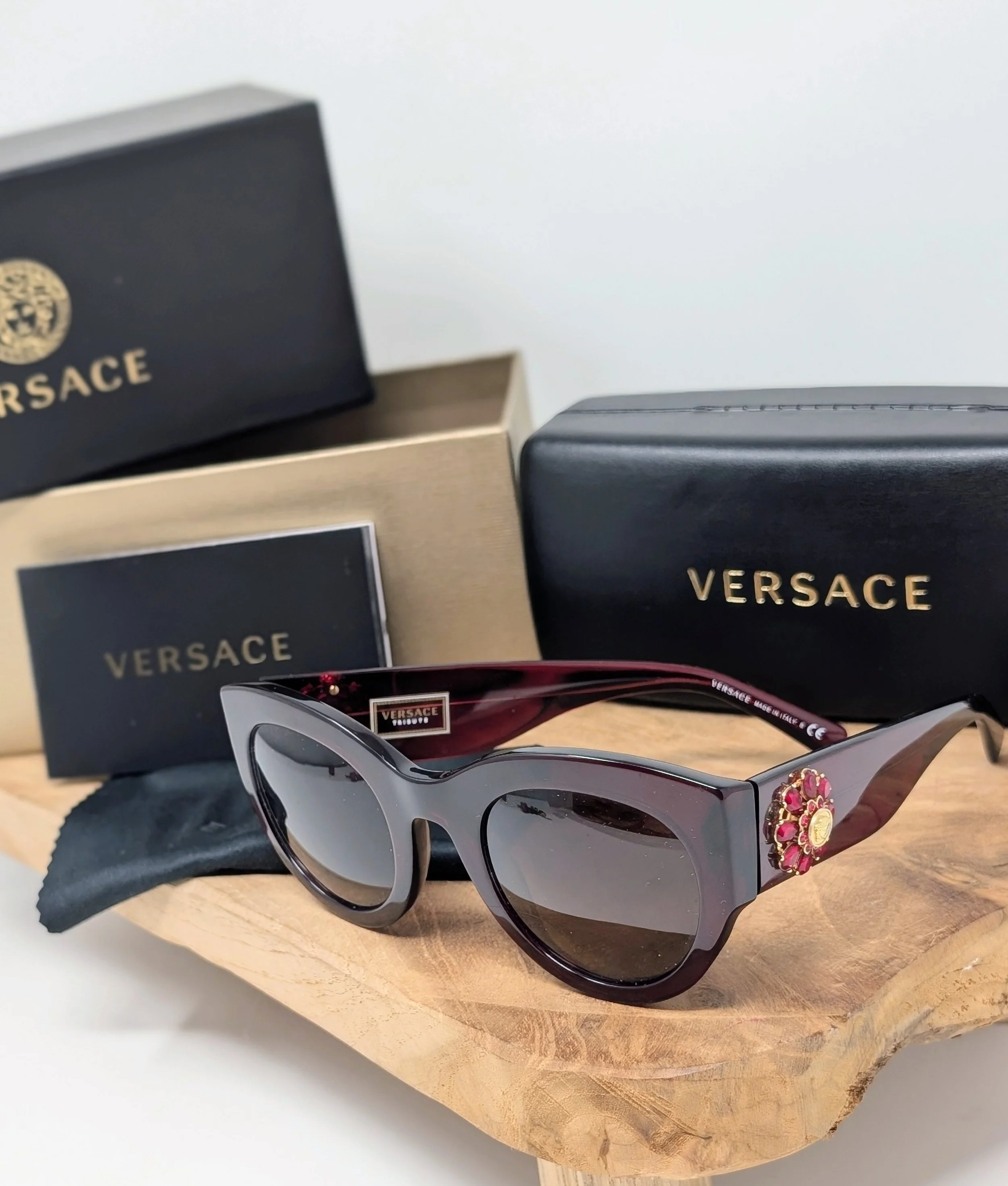 Eine Sonnenbrille mit dunklen Gläsern und einem Verasace-Logo, die auf einem Holztablett liegt, neben zwei Verpackungen mit Verasace-Designs. Vintage, Designer Brille