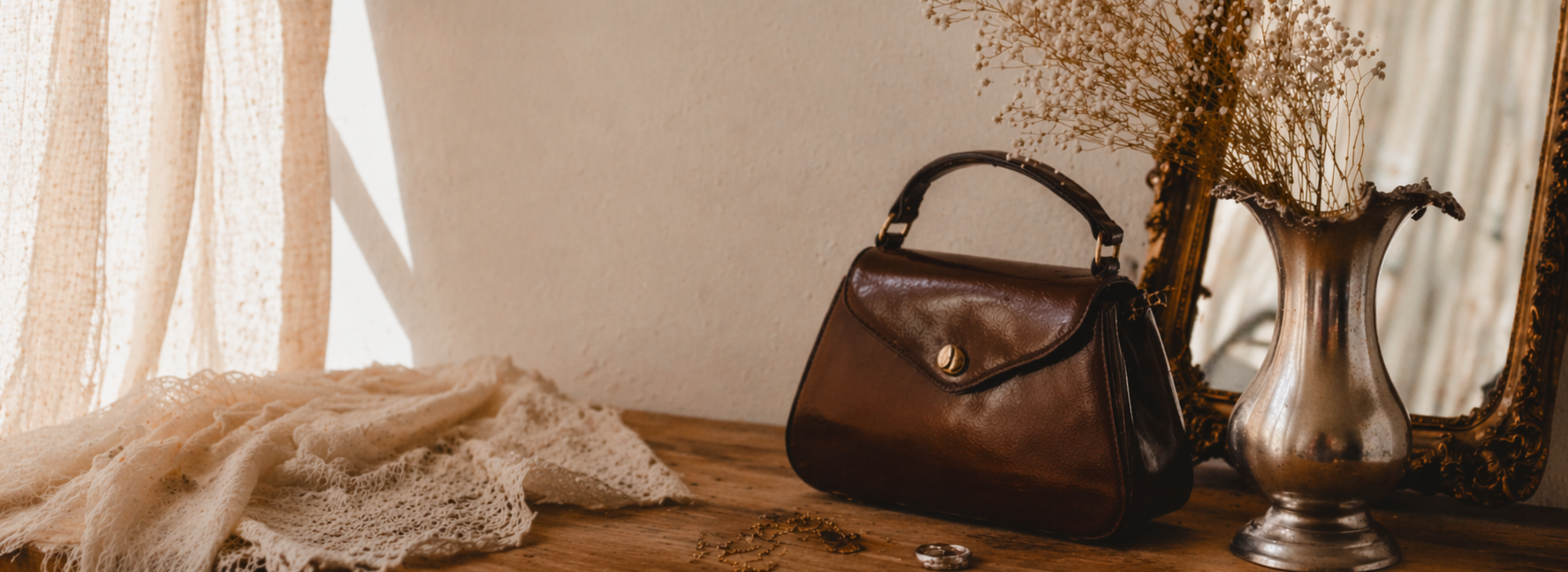 Ein brauner Ledertasche auf einem Holzregal neben einem silbernen Vasen mit getrockneten Blumen und einem goldenen Bilderrahmen, beleuchtet von natürlichem Licht durch einen Vorhang.