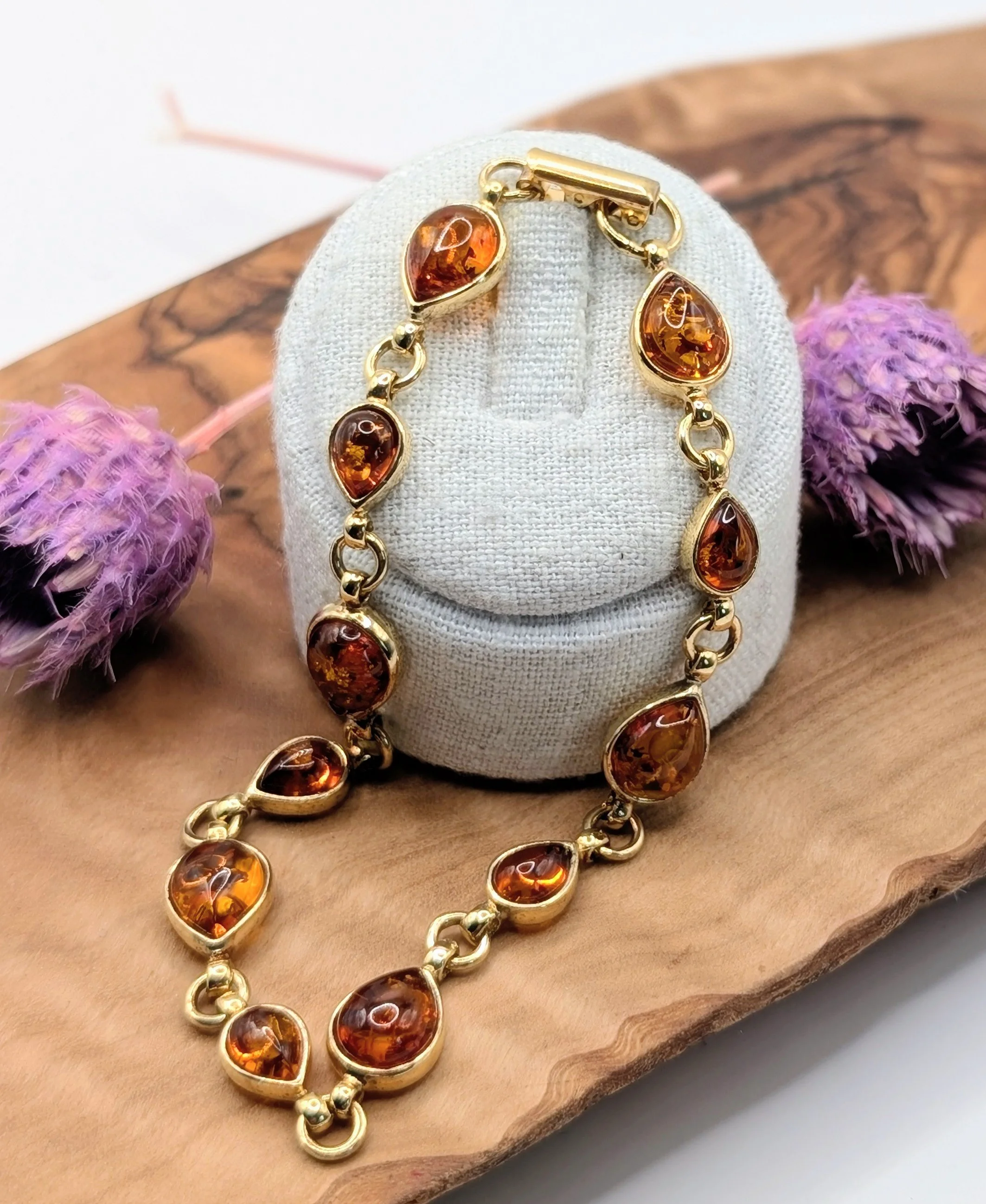 Goldene Kette mit amberfarbenen Edelsteinen, auf einem Holzstück mit lila Blumen im Hintergrund. Berstein Edelsteine, 925 silber Armkette, vergoldet.
