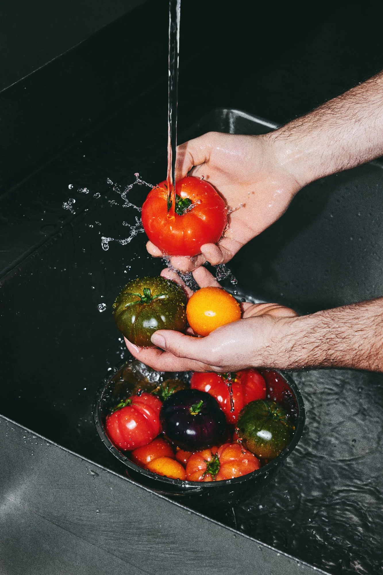 Hands washing colourful fresh tomatoes under running water in a kitchen sink.