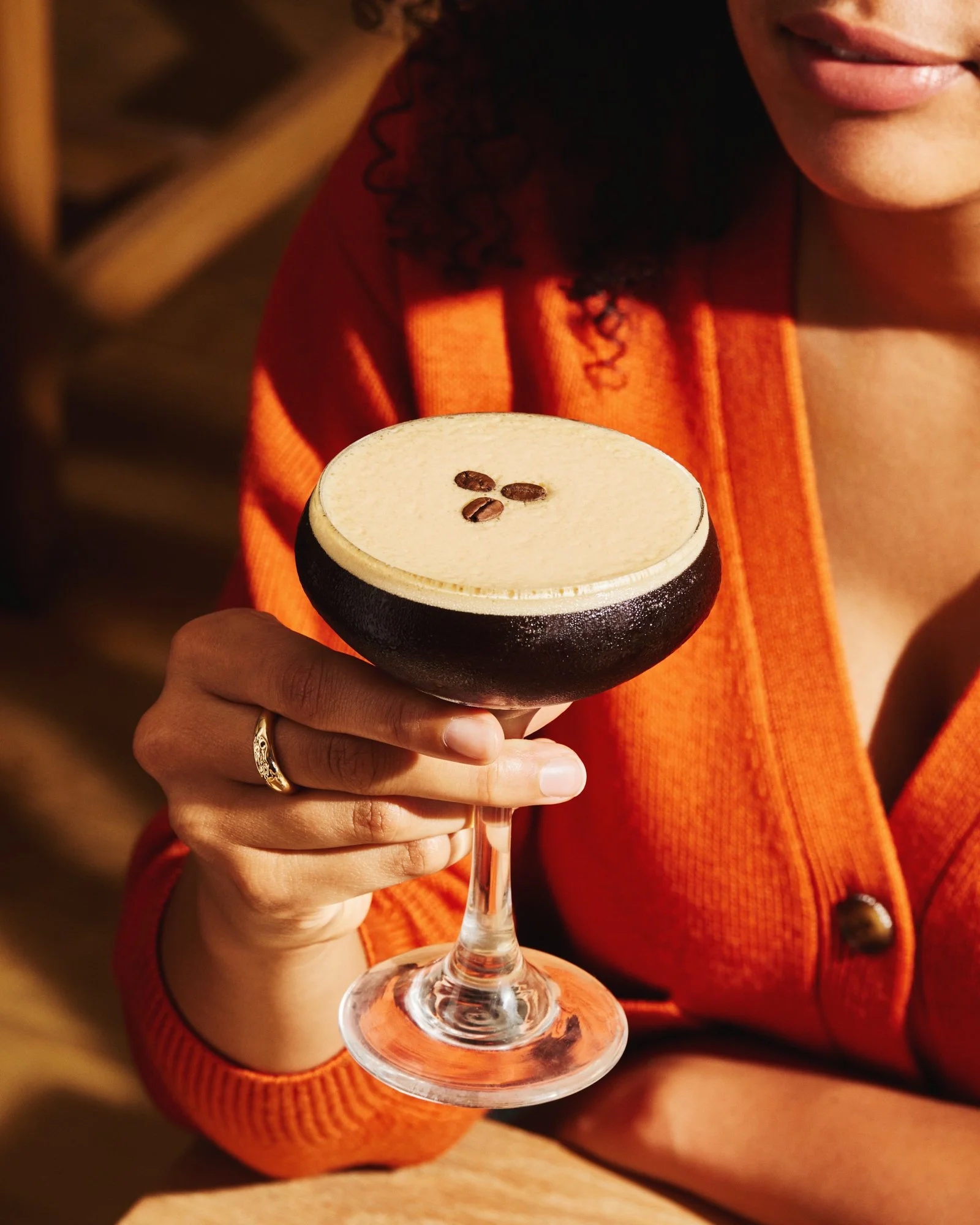 Woman in an orange cardigan holding an espresso martini in warm restaurant lighting.