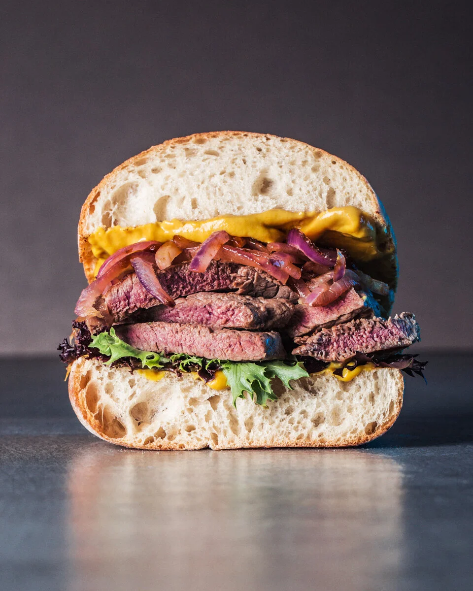 Stacked steak sandwich with sliced beef, caramelised onions and sauce against a dark background.