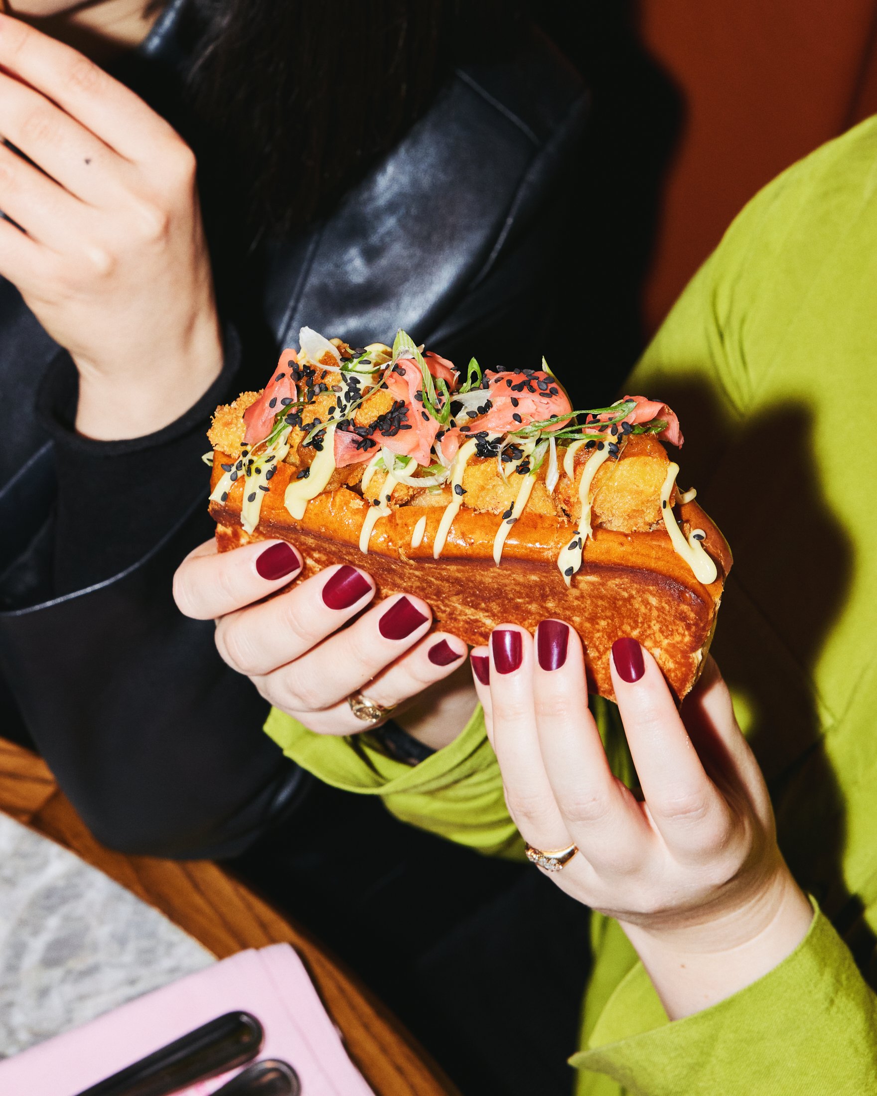 Hands holding a loaded lobster roll topped with colourful vegetables and sauces.