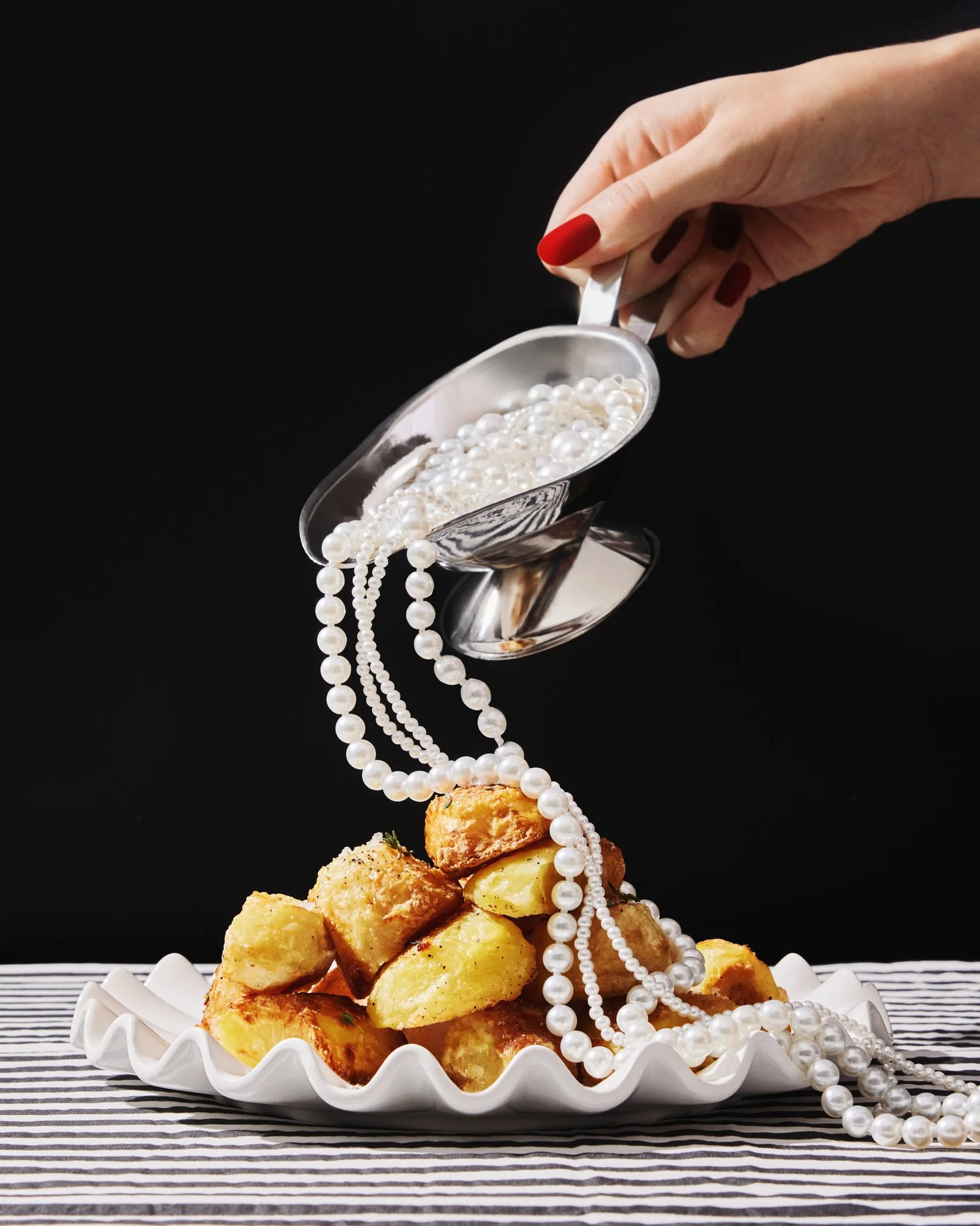 Surreal food photograph of pearls spilling from a silver gravy boat onto roast potatoes against a black background.