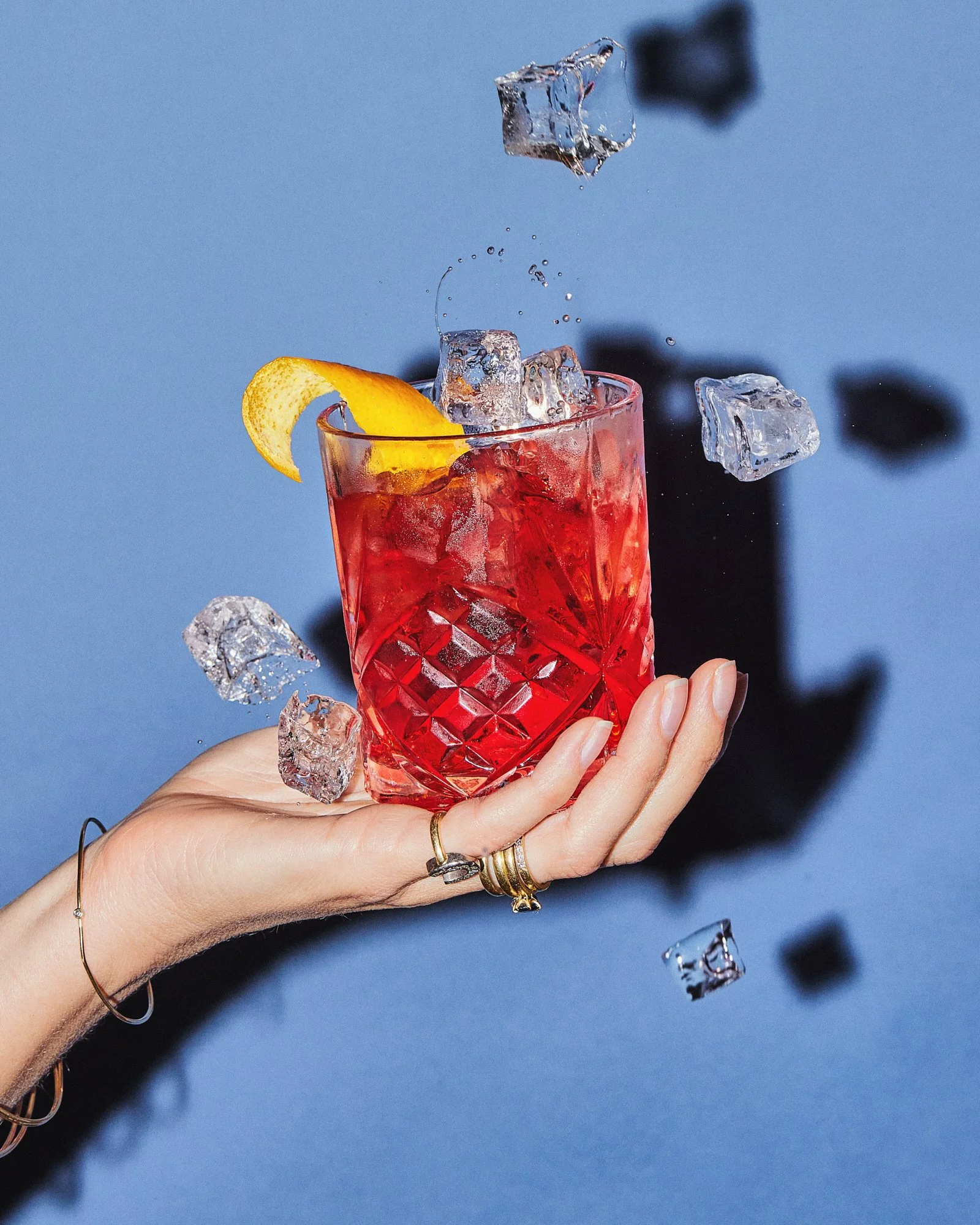 Hand holding a red cocktail as ice cubes fall around the glass against a blue background.