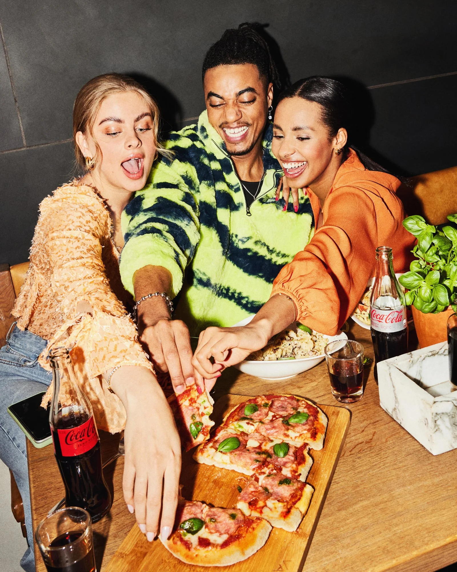 Three friends reaching for pizza together at a lively restaurant table.