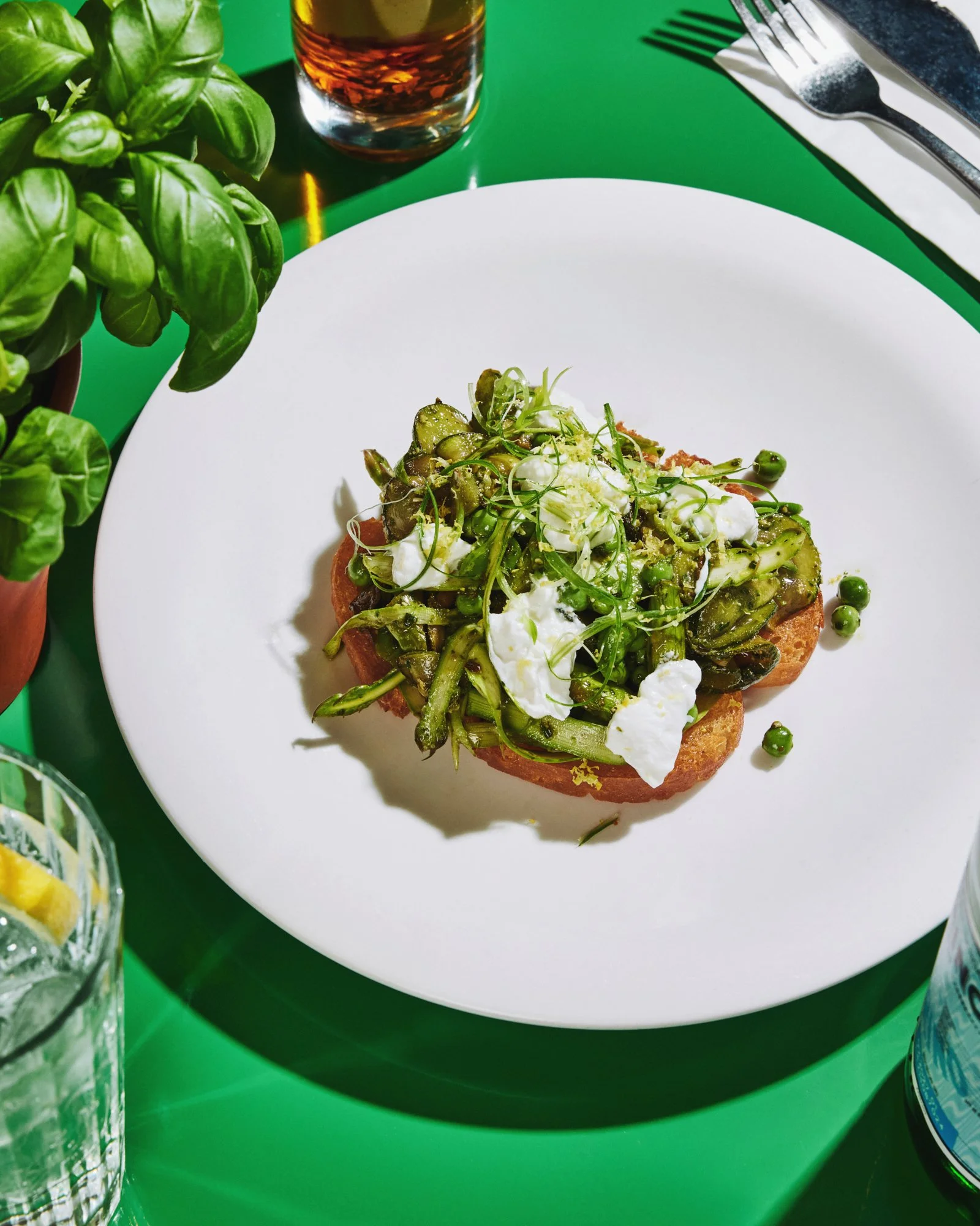 Overhead food photograph of a green vegetable bruschetta dish on a white plate surrounded by fresh basil.