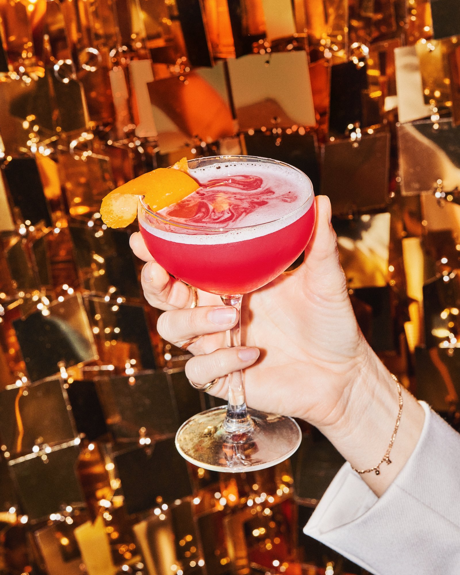 Hand holding a pink cocktail garnished with dried citrus against warm golden lights.