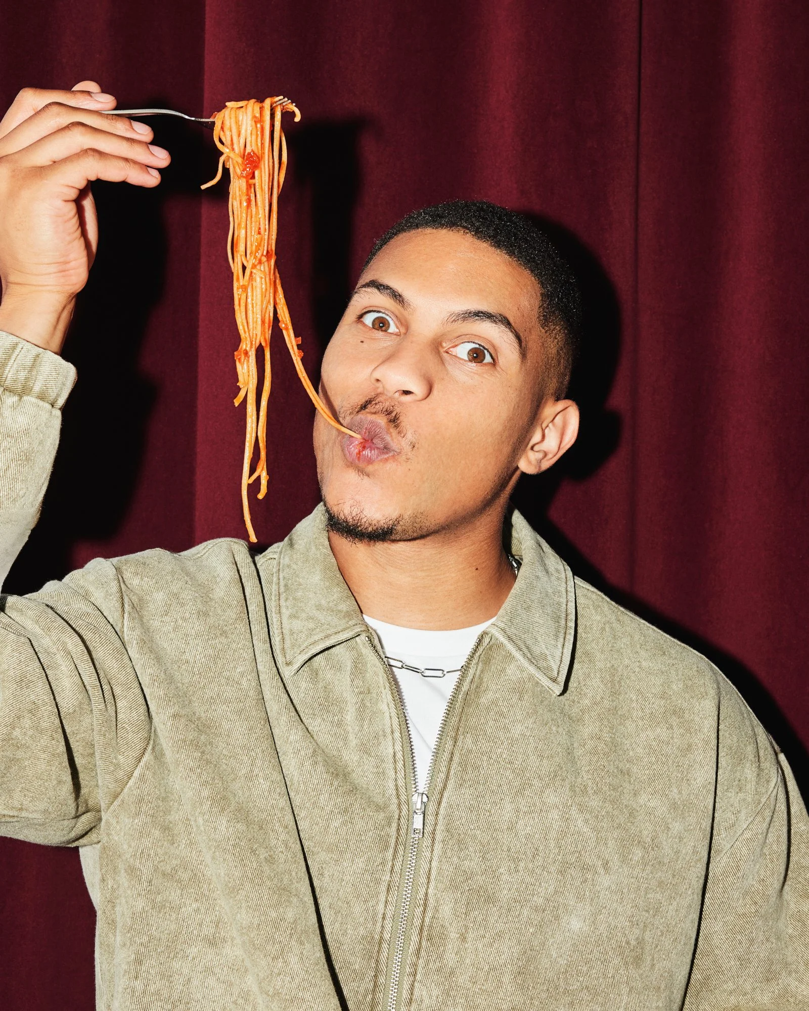 Man lifting a forkful of spaghetti above his mouth against a burgundy backdrop.