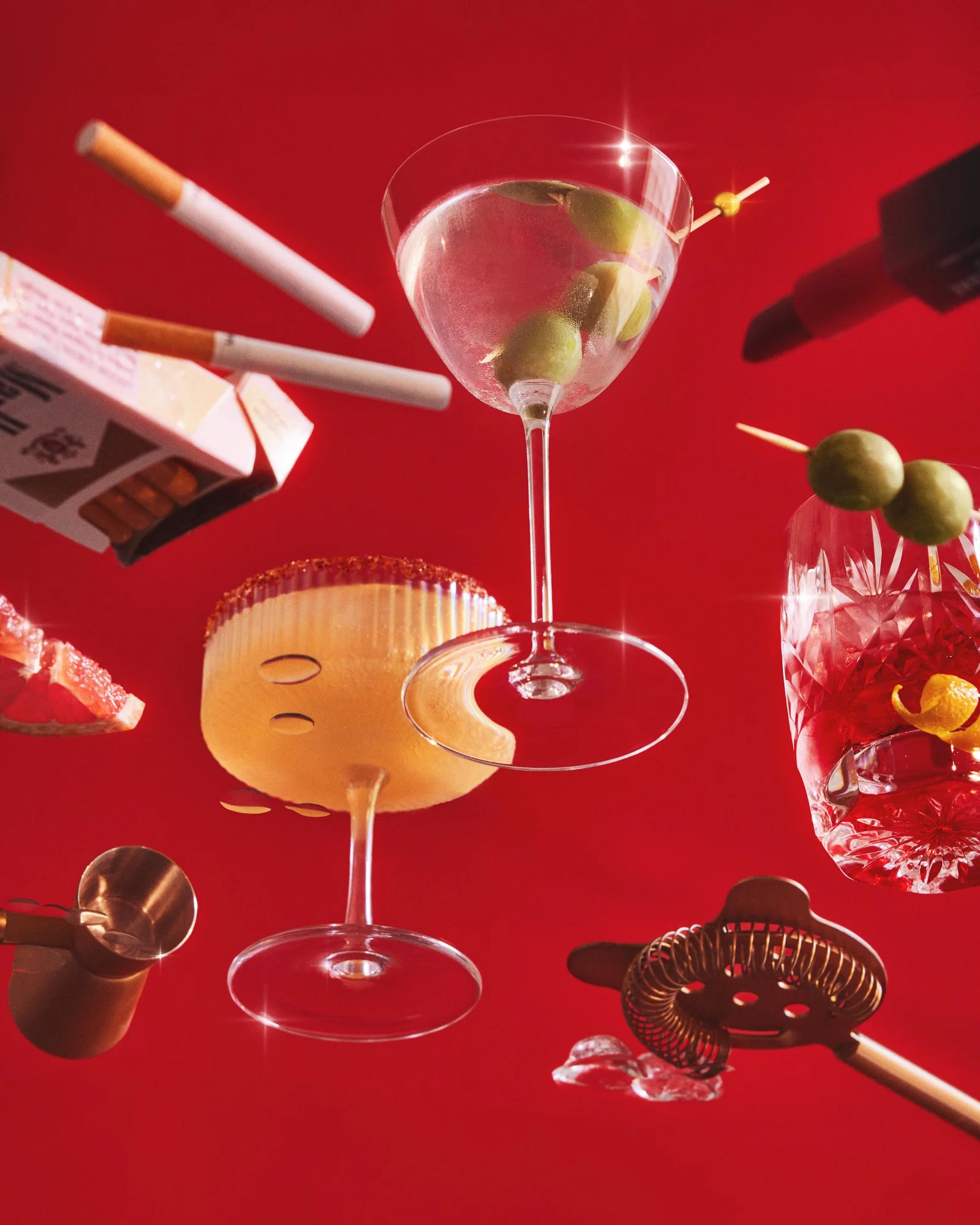Creative studio photograph of cocktails, olives and bar accessories against a vivid red background.