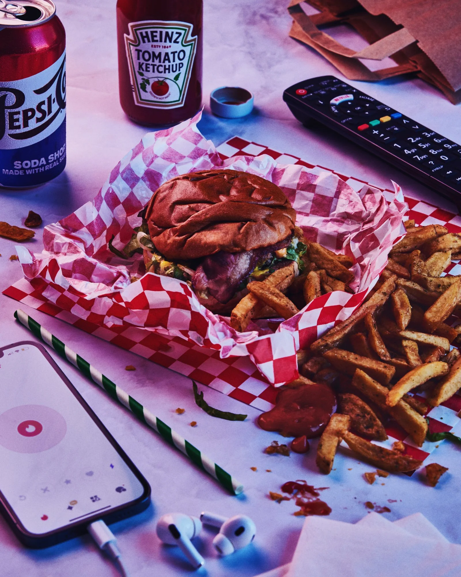 Burger and seasoned fries served on checked paper beside Pepsi, ketchup and a television remote.