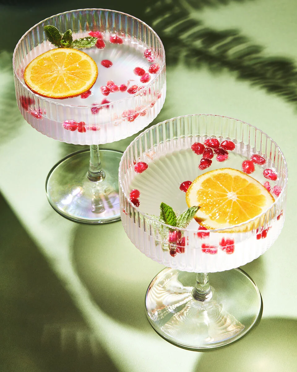 Two sparkling cocktails garnished with orange slices, pomegranate seeds and mint in ribbed glassware.