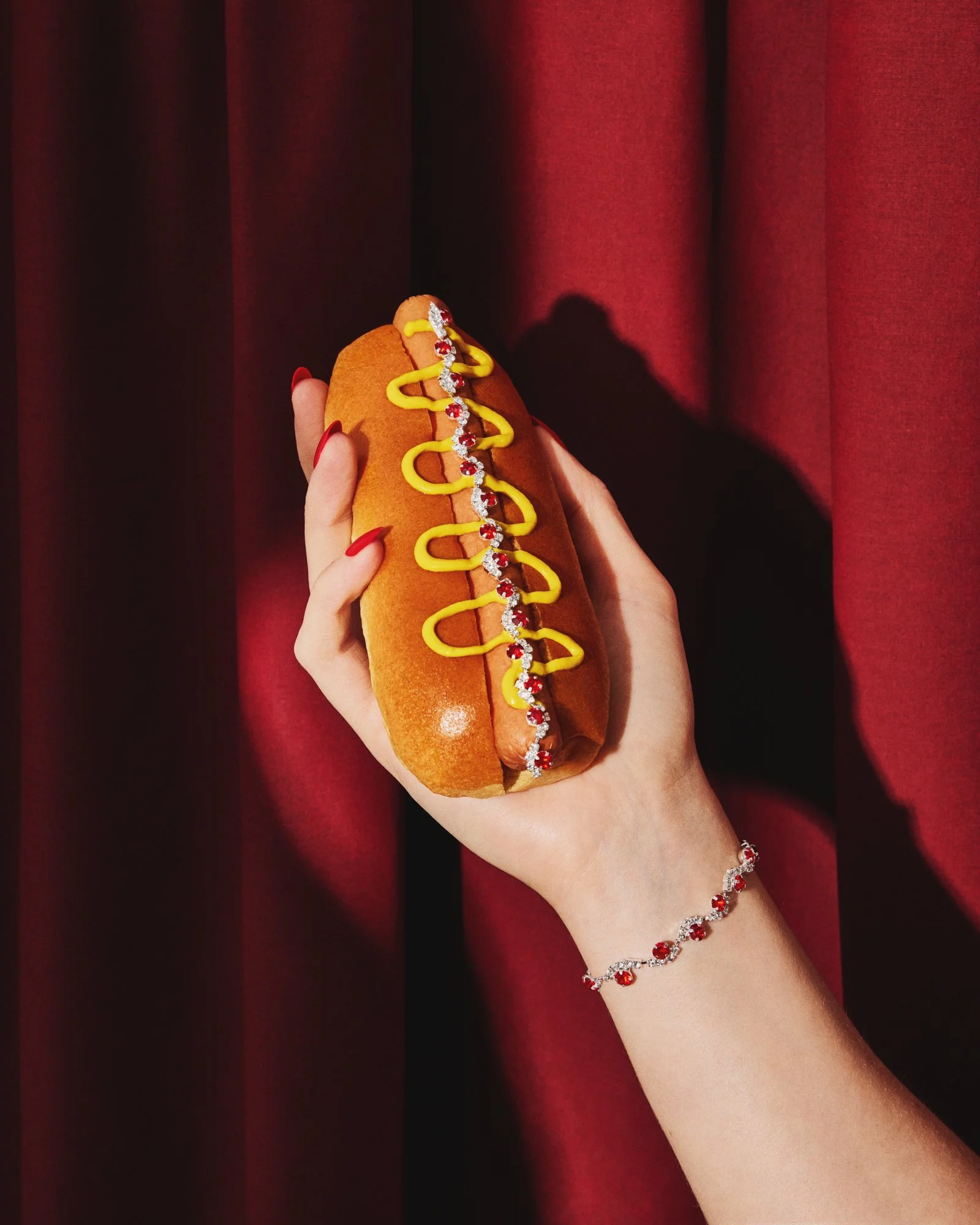 Hand holding a hot dog topped with mustard and jewellery against a deep red curtain backdrop.