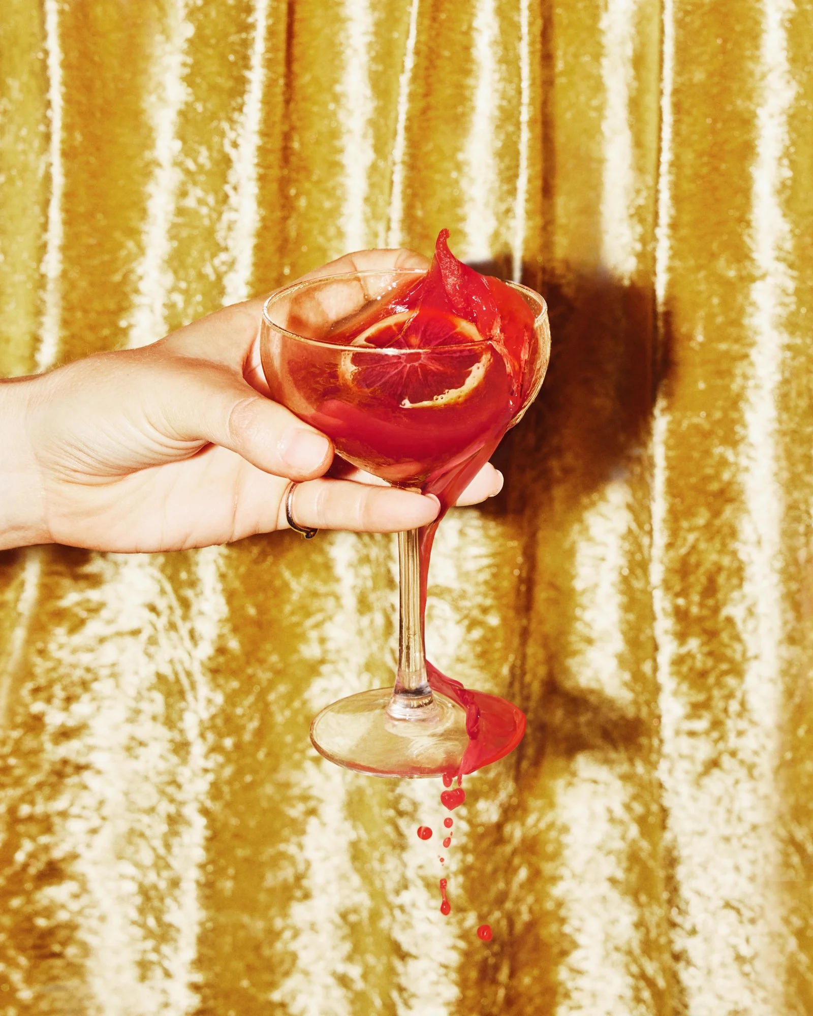 Hand holding a red cocktail in a coupe glass against a gold velvet curtain.