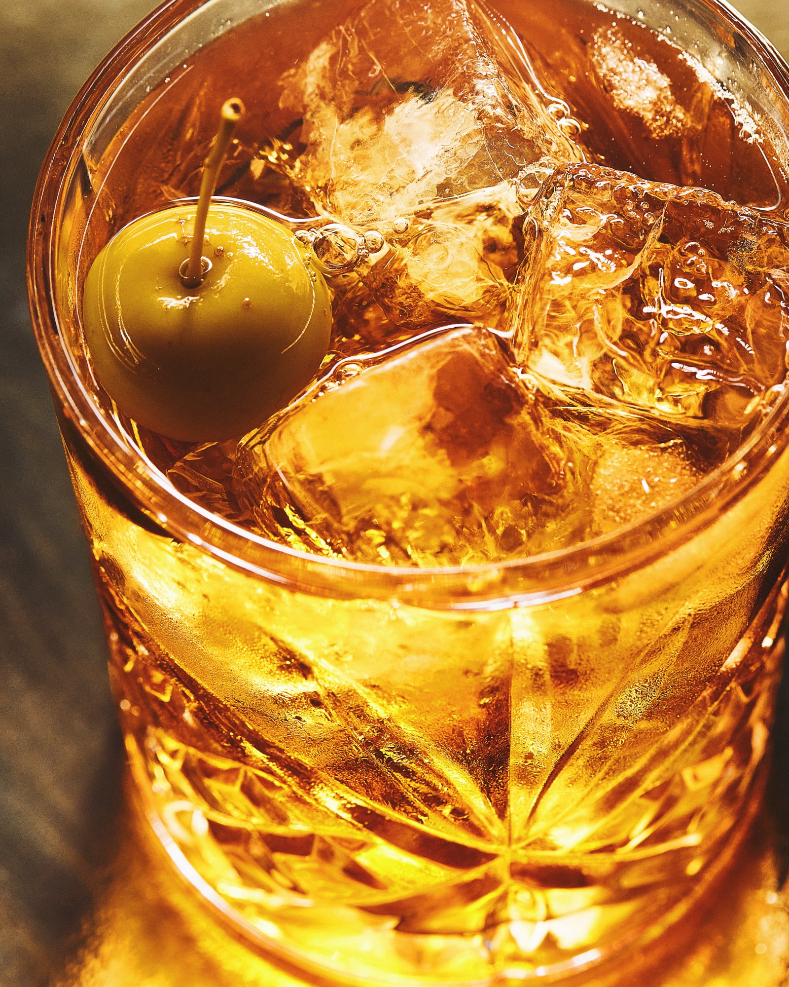 Close-up of an amber cocktail with ice and a green olive in a cut-glass tumbler.
