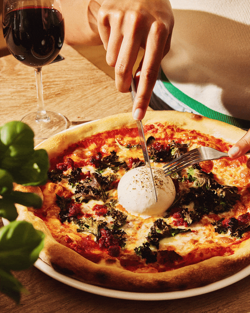 Stop motion GIF of pizza topped with burrata, vegetables and herbs being cut at a restaurant table.