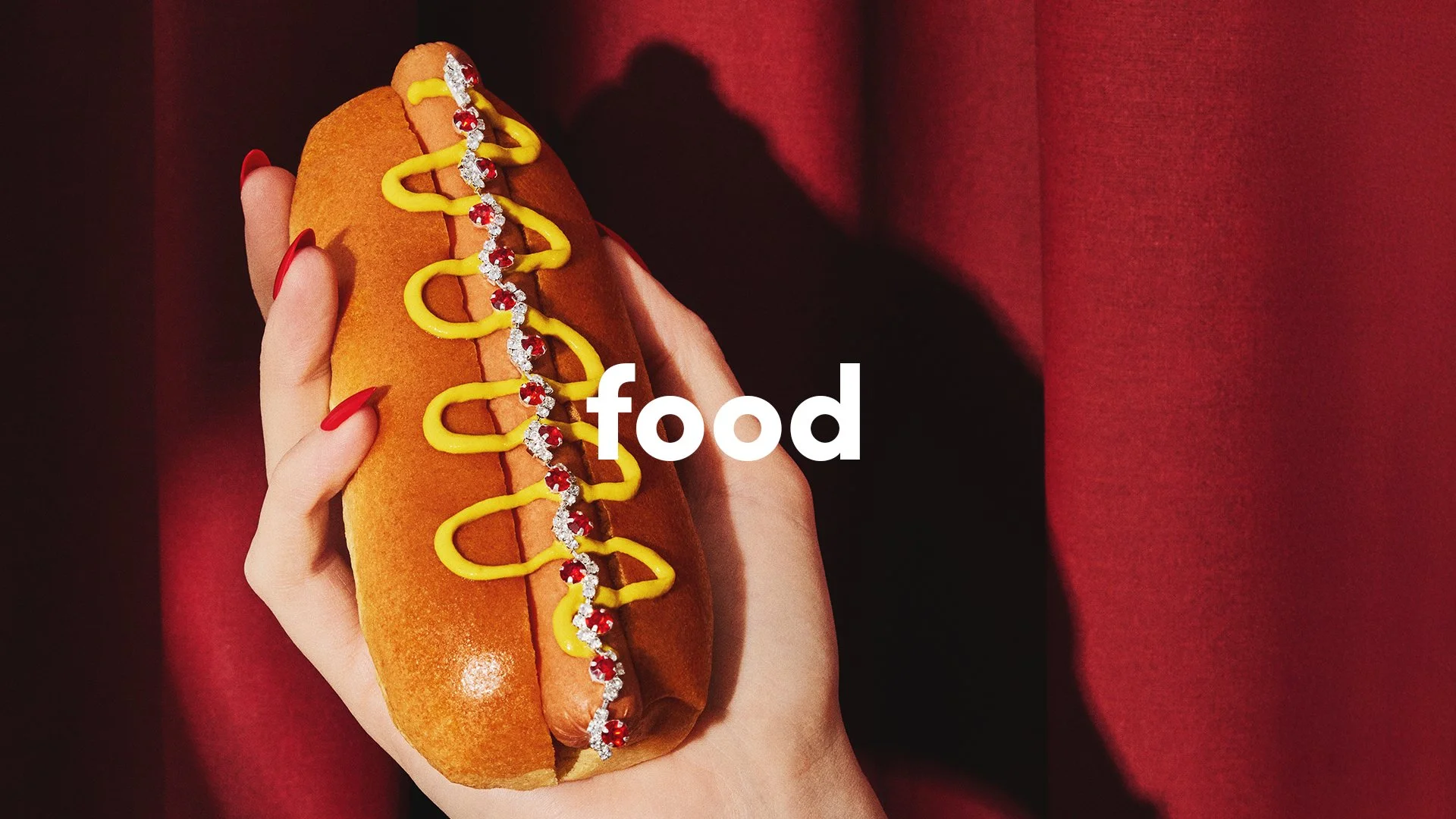Hands holding a hot dog topped with mustard and jeweled embellishments against a red backdrop overlaid with the word “food.”
