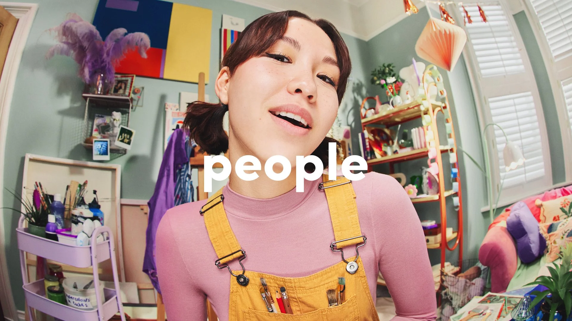 Smiling artist wearing yellow overalls and a pink shirt in a colorful art studio overlaid with the word 'people'