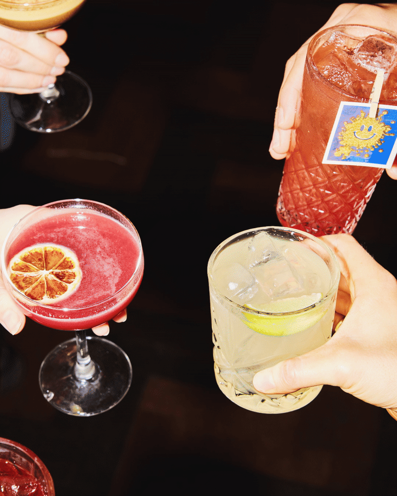 Stop motion GIF of friends raising colourful cocktails together, photographed from above.