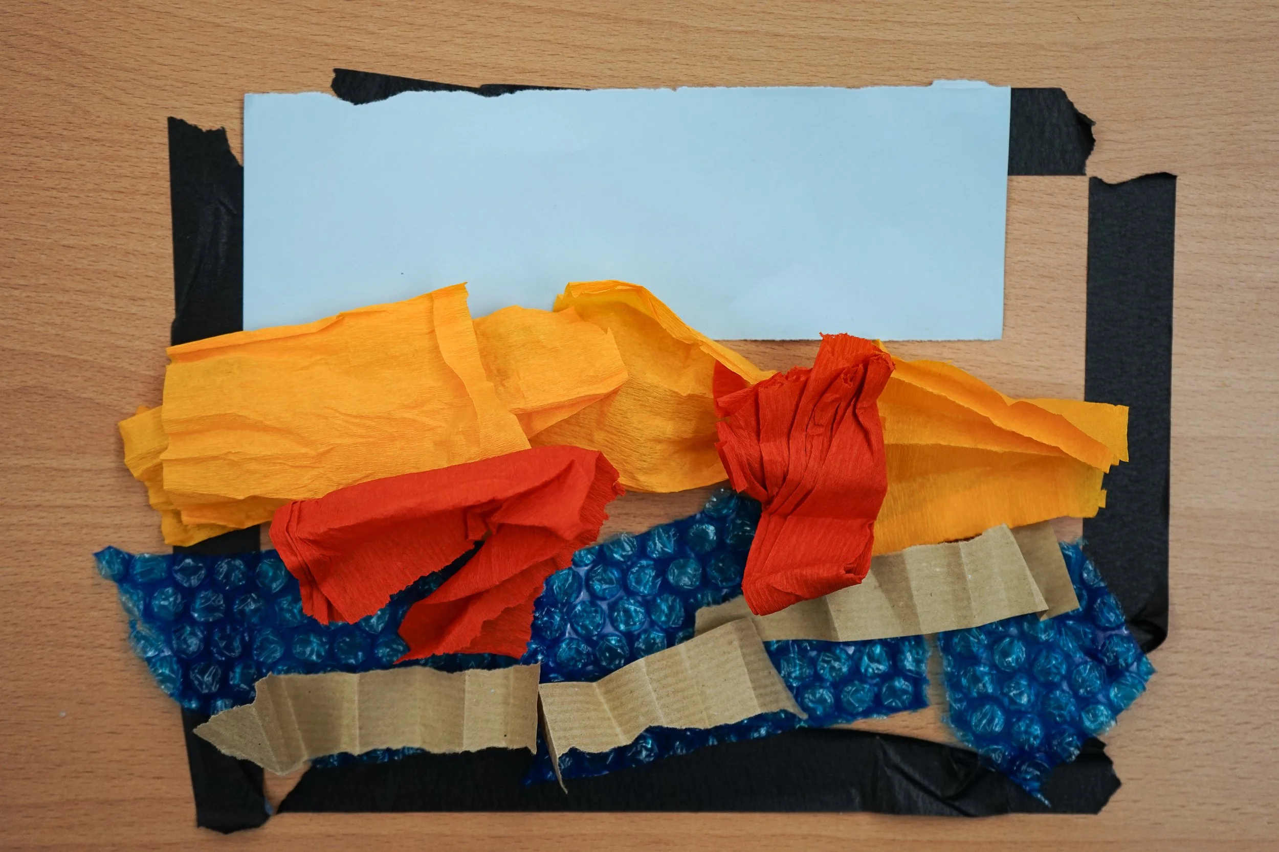 Colorful tissue paper pieces arranged on black foam padding, with bubble wrap and cardboard at the bottom, on a wooden surface.