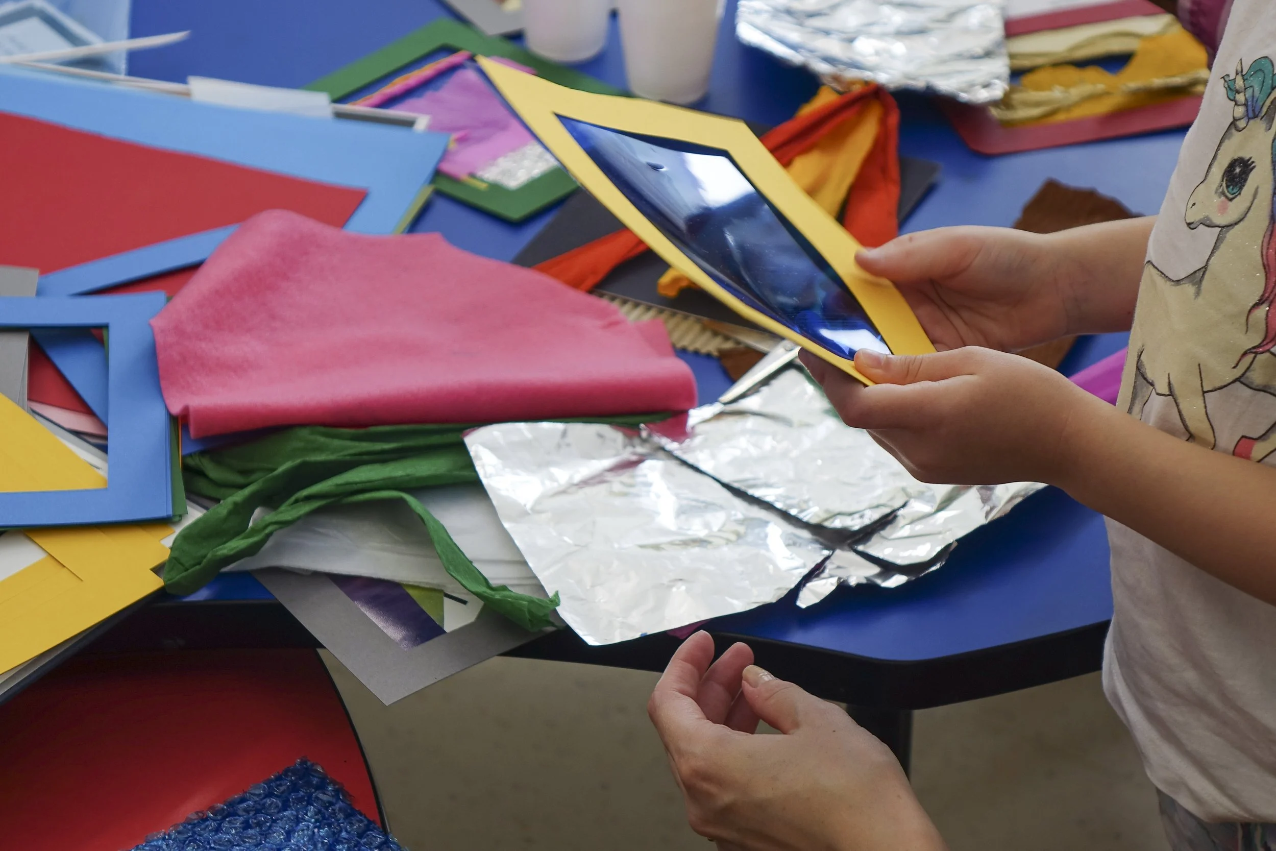 Child holding a colorful picture frame surrounded by craft supplies like foam sheets, tissue paper, and foil.