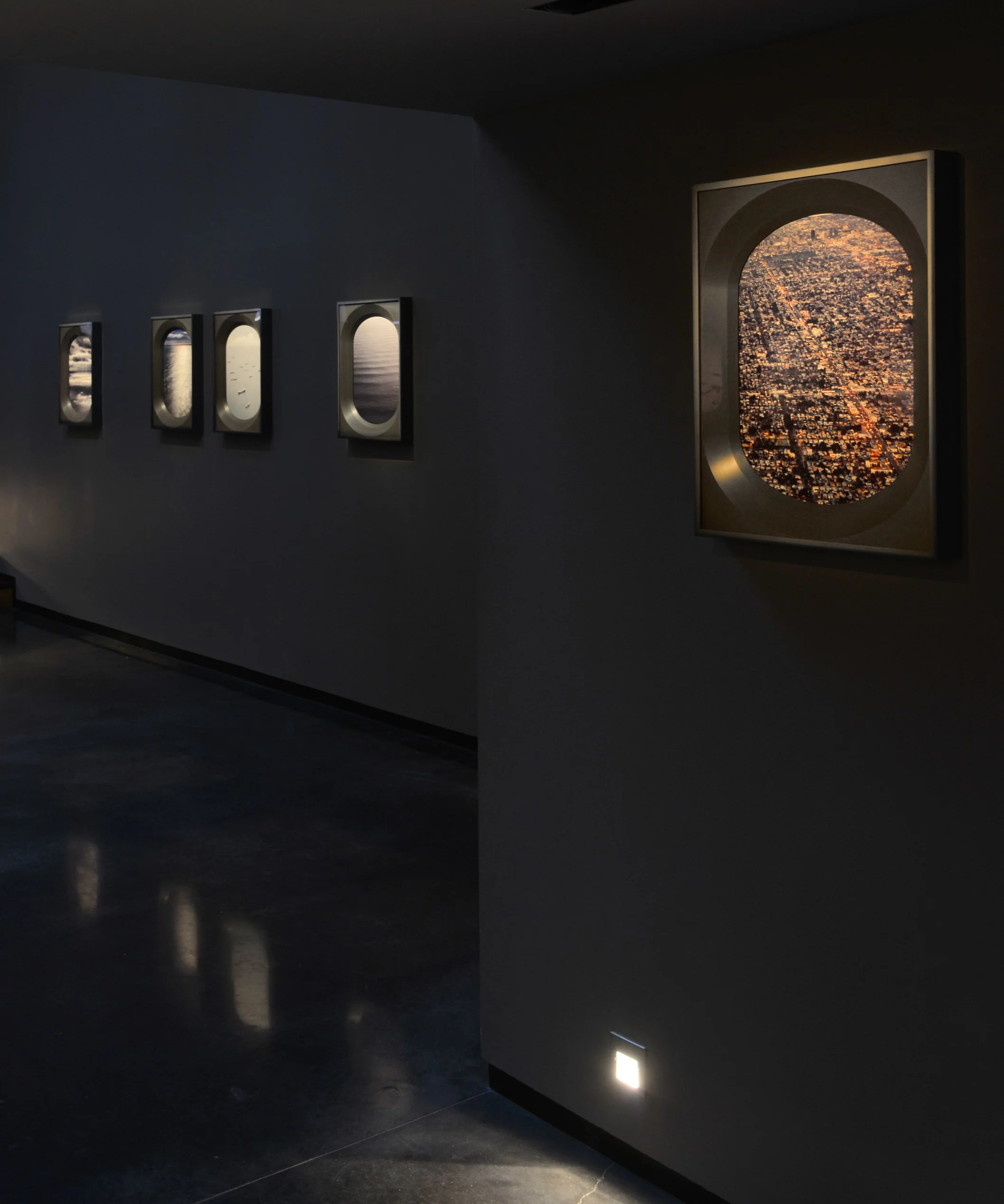 A dark museum or gallery interior with four small art frames on the left wall and a large window or screen on the right showing an aerial view of a city at sunset.