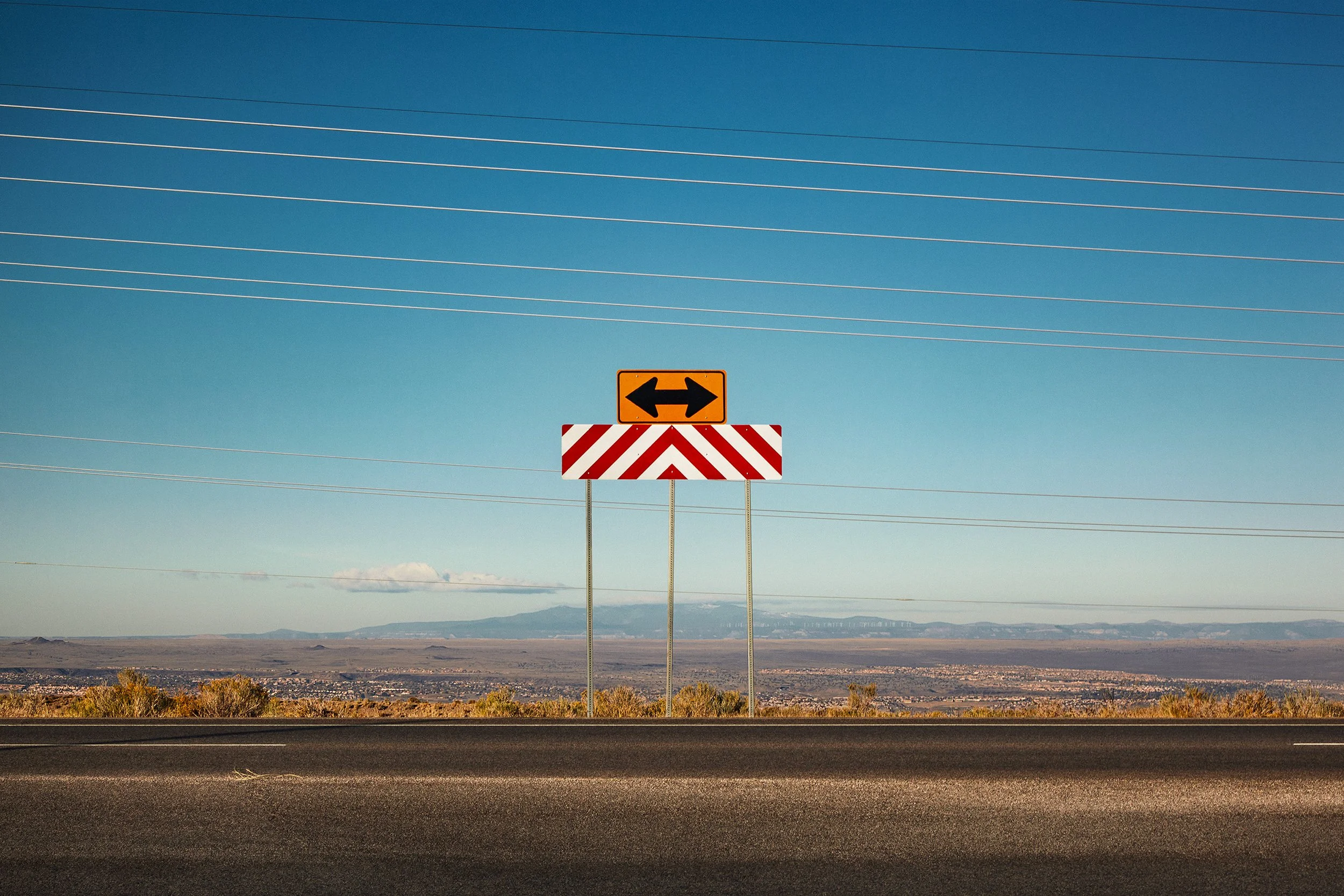 A highway with a directional sign indicating both left and right turns, set against a background of mountains and a clear blue sky.