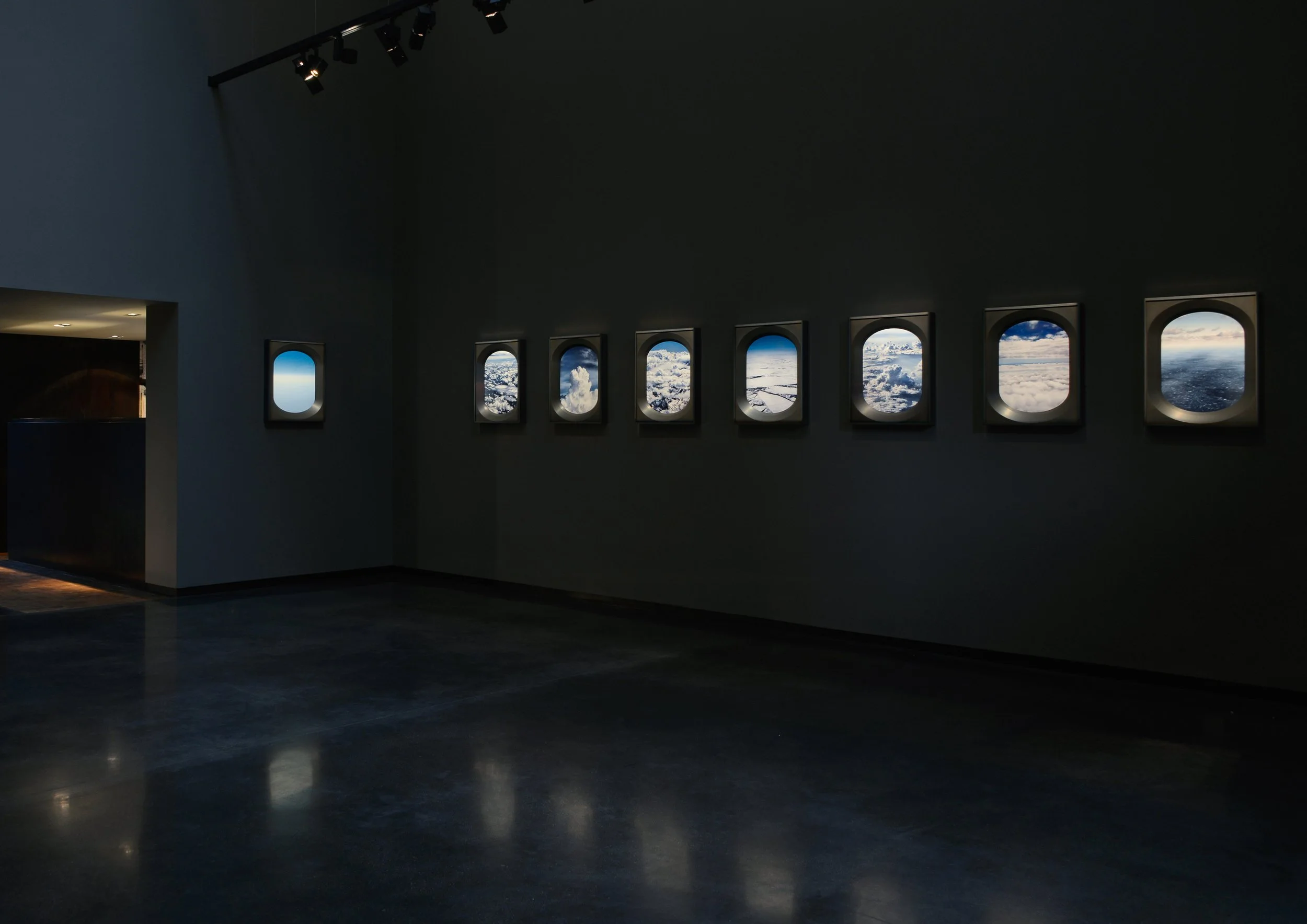 An interior space with a dark wall featuring a row of small airplane windows showing clouds and sky outside. The room has a dark floor with reflections and a doorway leading to a lit area.