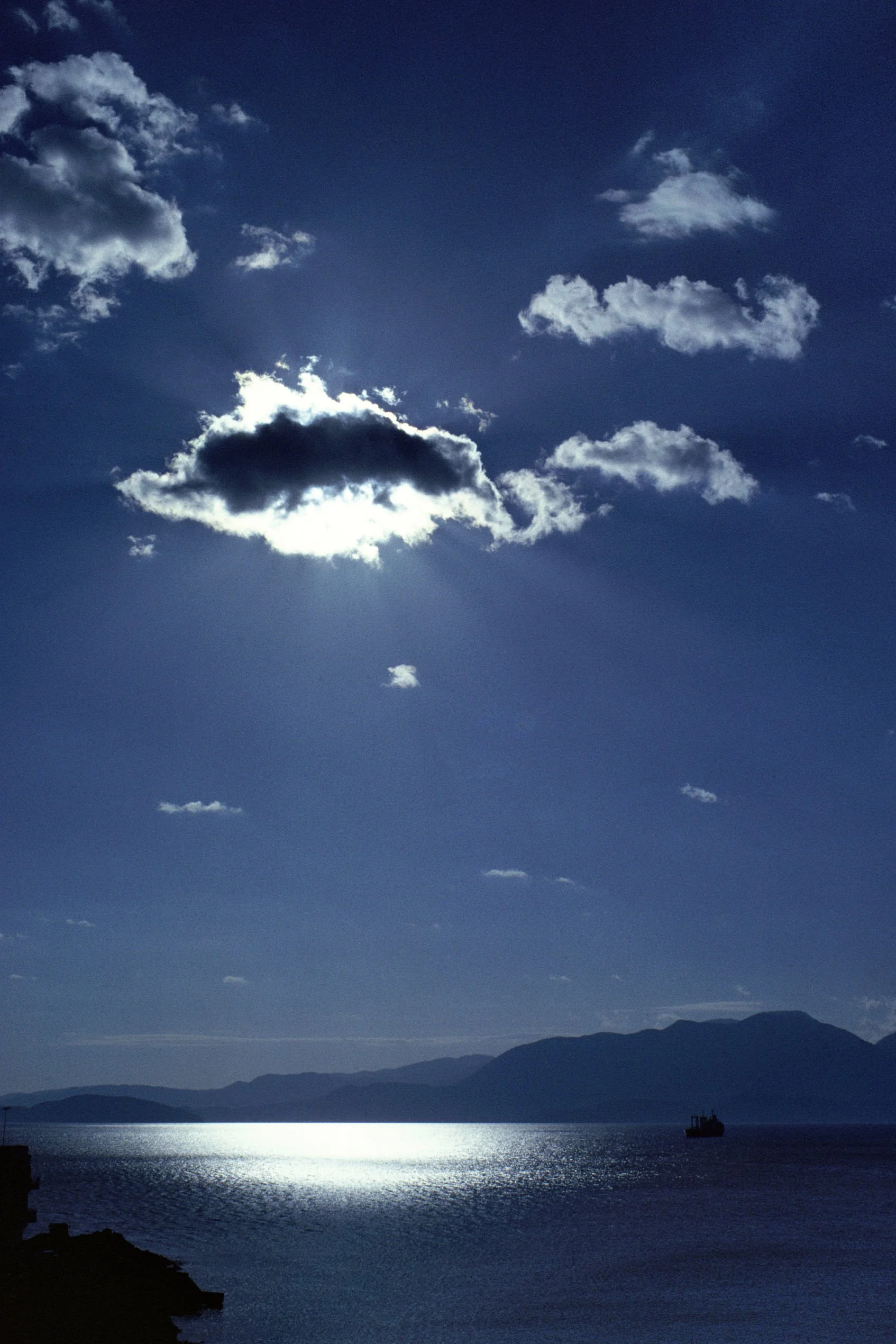 Sun partially hidden behind clouds over a body of water with mountains in the background and a boat on the water, silhouetted dark rocks in the foreground.