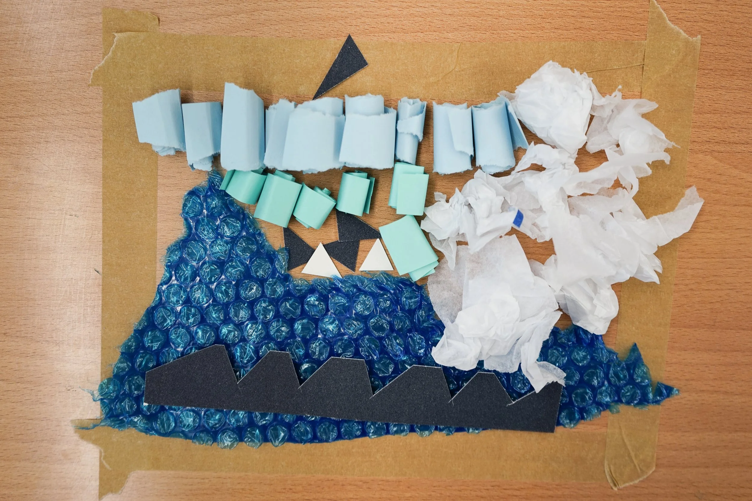Collage of textured craft materials including blue bubble wrap, black foam, light blue foam, white tissue paper, and black cutouts, arranged on a wooden surface lined with masking tape.