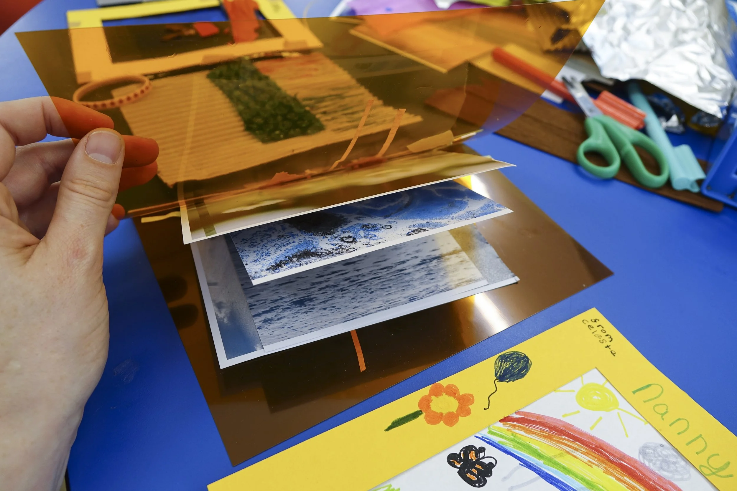 A person holding a transparent orange sheet over printed photographs on a table. The table has arts and crafts supplies including scissors, markers, and papers. A colorful children's drawing with flowers, sun, and balloon is visible.