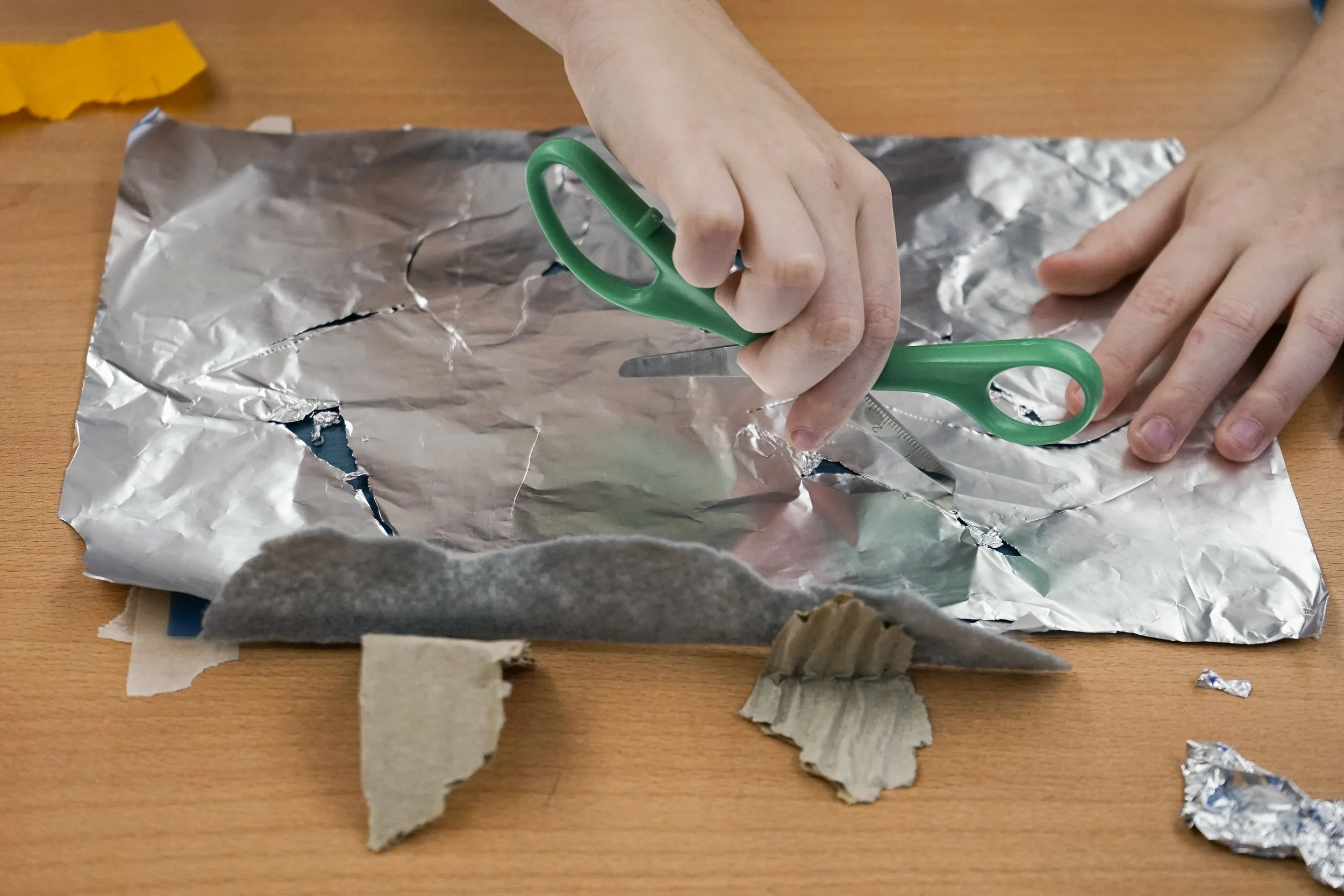 Person cutting aluminum foil with green-handled scissors on a wooden table, surrounded by torn pieces of cardboard and foil