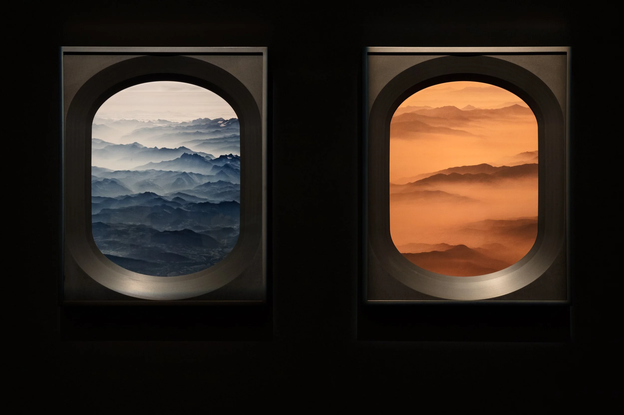 View from airplane windows showing mountain ranges, with the left window displaying a blue-toned mountain landscape and the right window showing an orange-toned mountain landscape at sunset or sunrise.