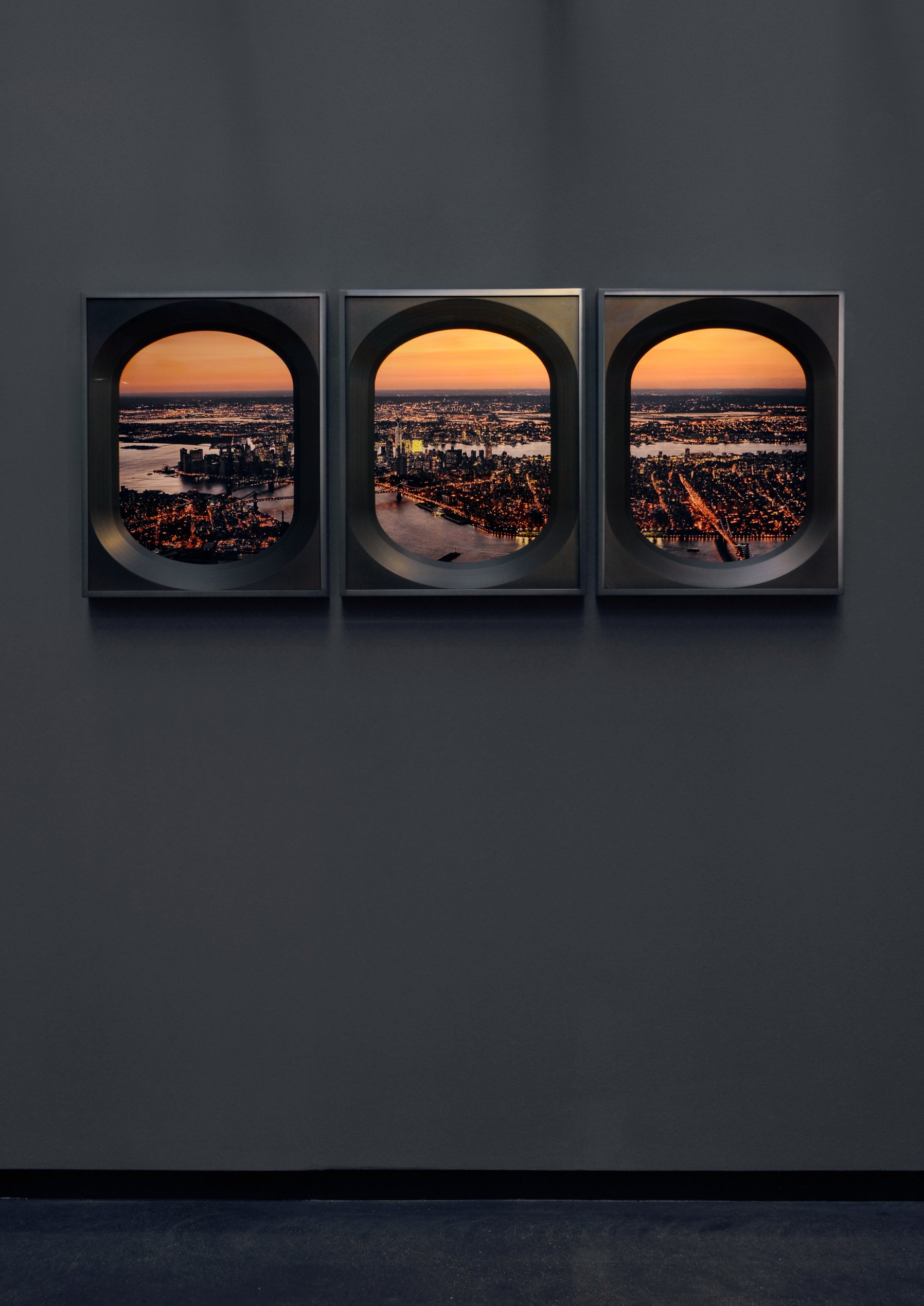Three framed pictures of a cityscape at sunset, viewed through airplane windows, mounted on a plain gray wall.