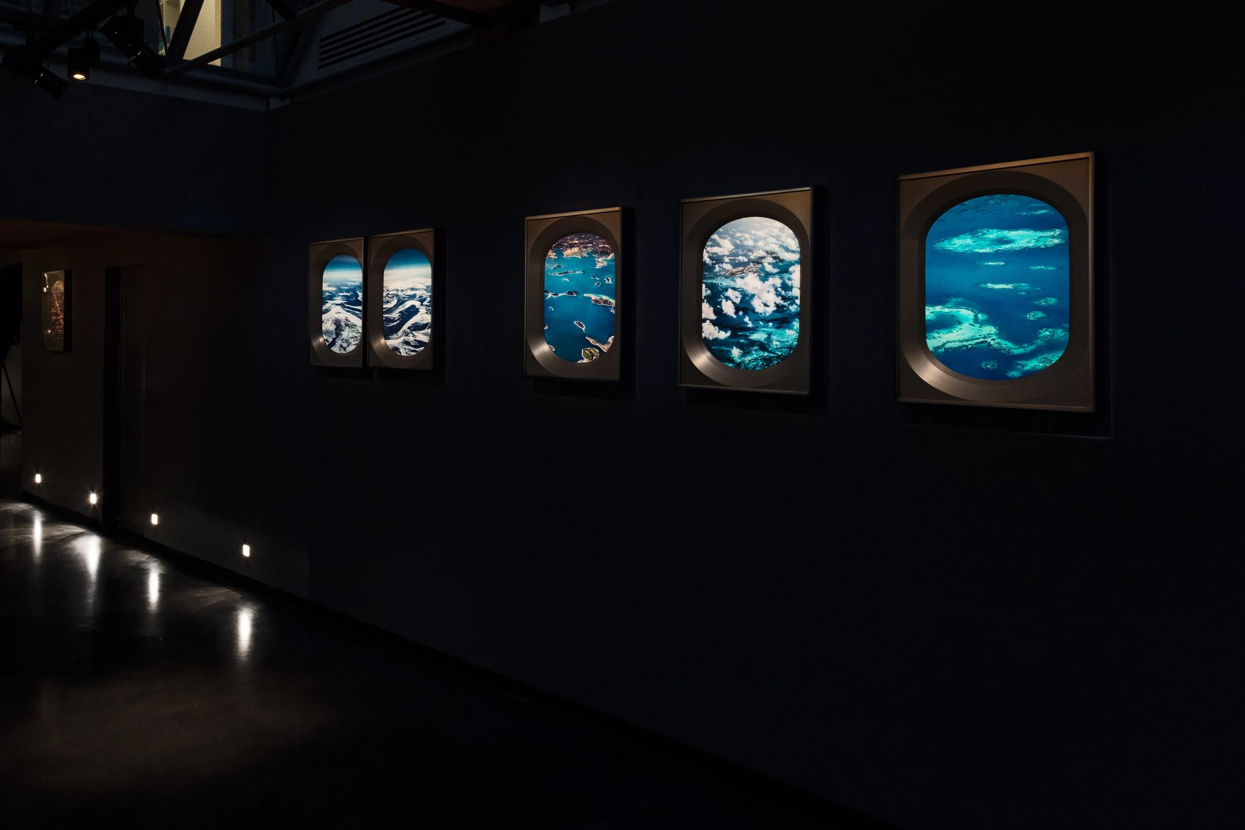 Interior of an exhibit with framed aerial photographs of landscapes through airplane windows, displayed on a dark wall.