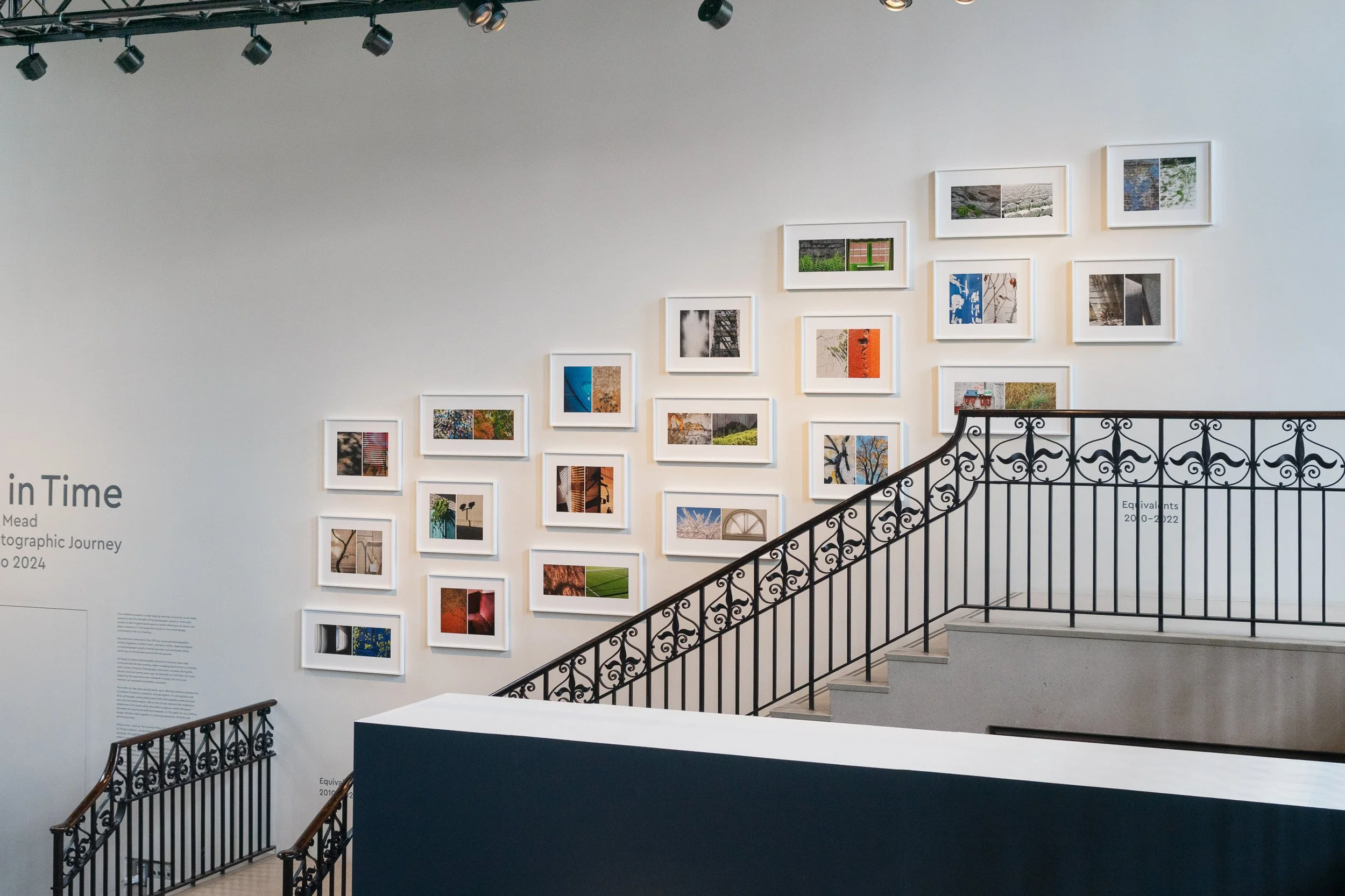 Gallery wall with multiple framed photographs in an art gallery, adjacent to a staircase with decorative black railing.